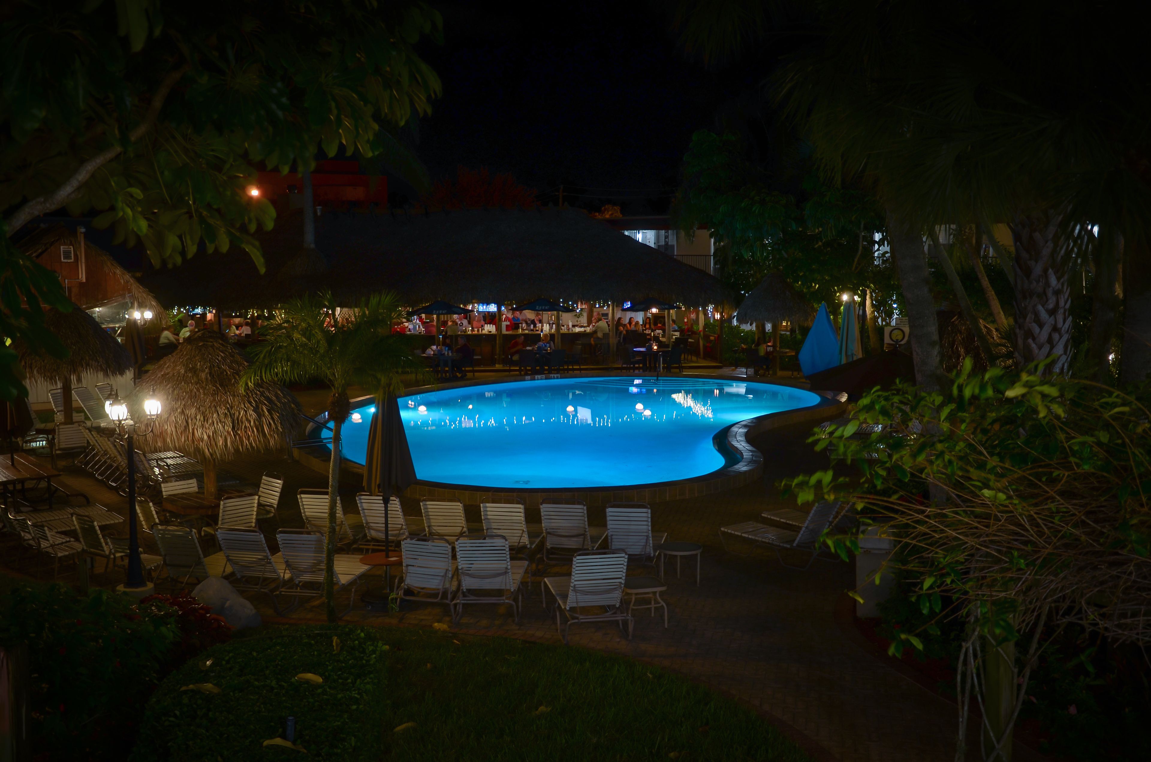 A nighttime swimming pool area with lounge chairs and a bar area in the background.