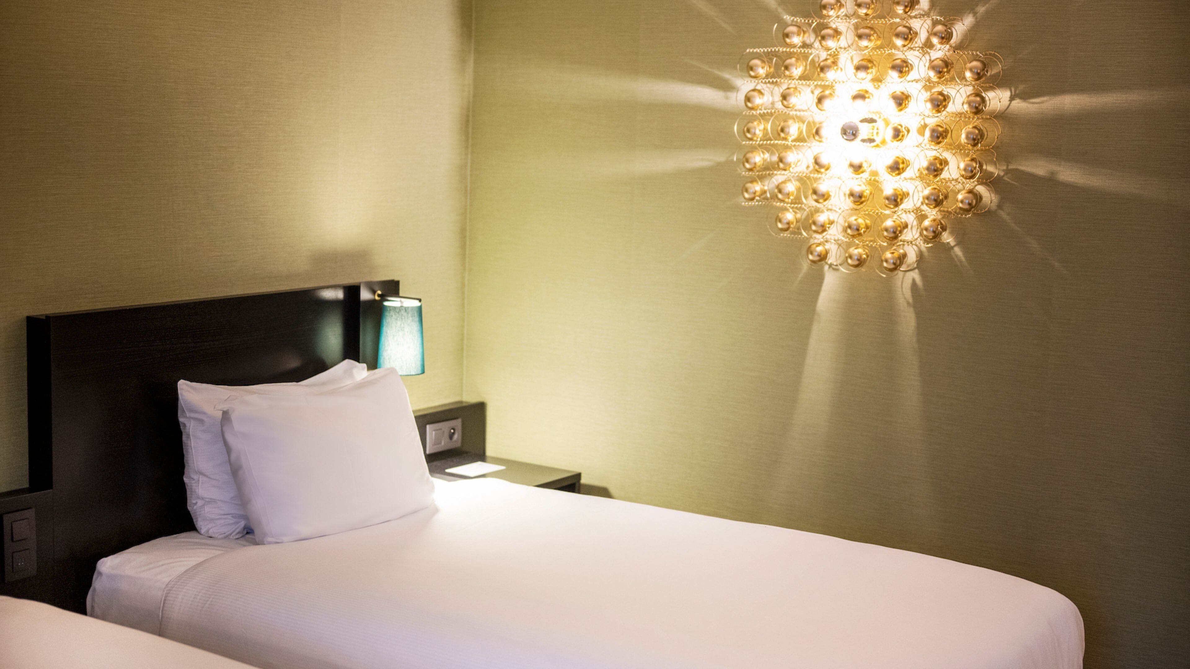 Een hotelkamer met een net opgemaakt bed, een nachtkastje met een lamp en een wandlamp.