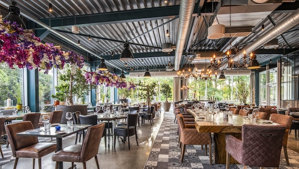 A spacious restaurant with high ceilings, exposed ductwork, and large windows offering views of greenery. Tables are set with wine glasses and plates, and the floor features a patterned tile design.