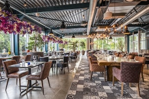 Un restaurante espacioso con techos altos, conductos expuestos y grandes ventanales que ofrecen vistas a zonas verdes. Las mesas están dispuestas con copas de vino y platos, y el suelo presenta un diseño de baldosas con motivos.