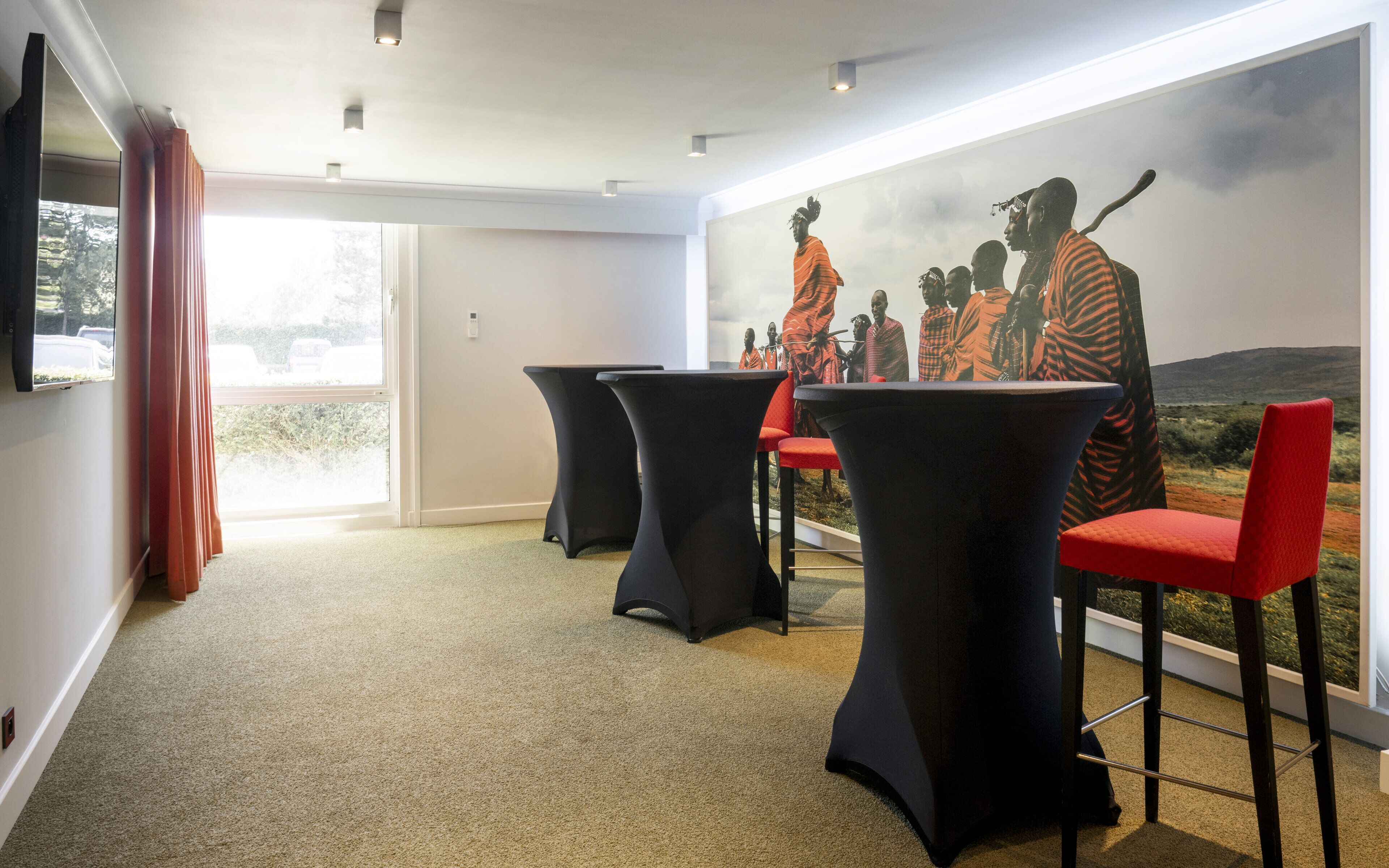 A room with a large window, a flat-screen TV, and a mural of a group of people. There are three black tables with red chairs.