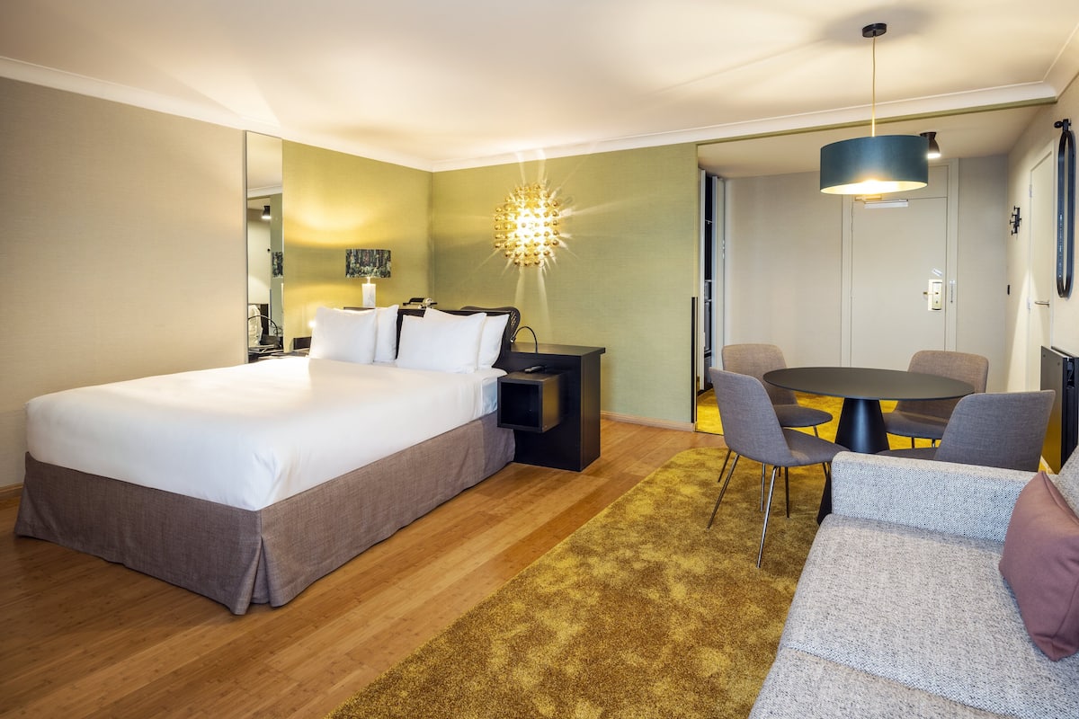 A hotel room with a large bed, a desk with a chair, a round table with chairs, and a sofa.