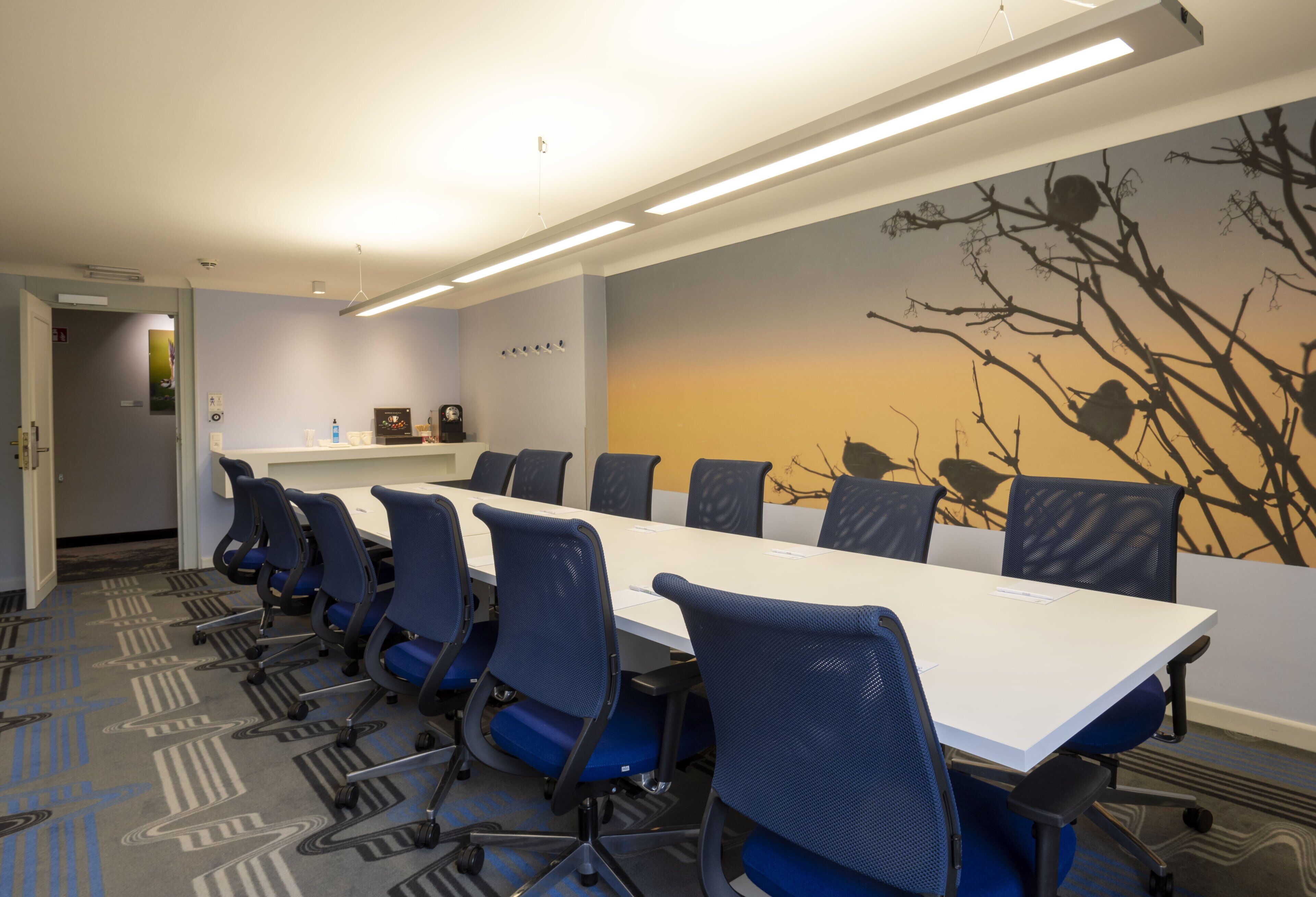 Une salle de conférence avec une longue table et des chaises bleues, une fresque représentant des branches d’arbres et un luminaire au plafond.