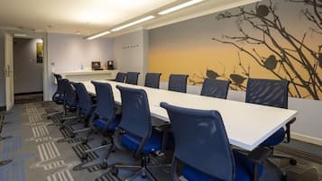 Une salle de conférence avec une longue table et des chaises bleues, une fresque représentant des branches d’arbres et un luminaire au plafond.