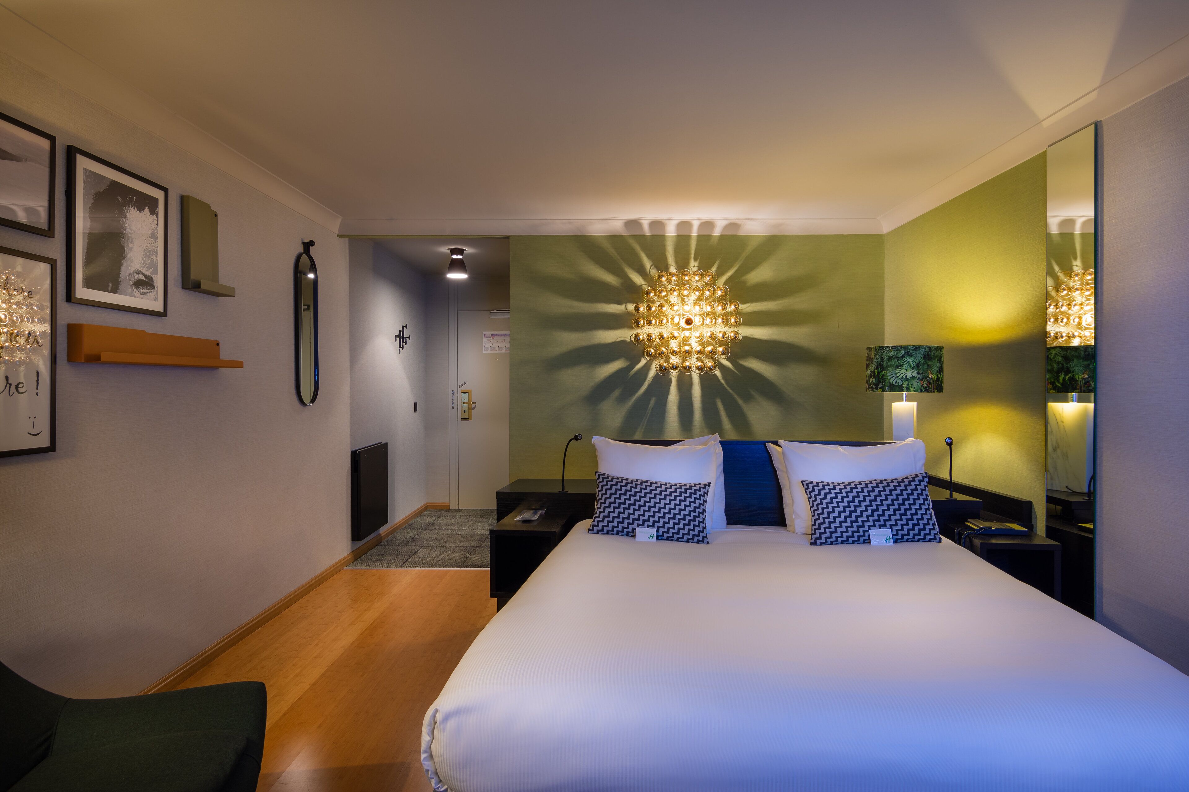 A hotel room with a large bed, bedside tables, a nightstand, a mirror, and a wall-mounted light fixture.
