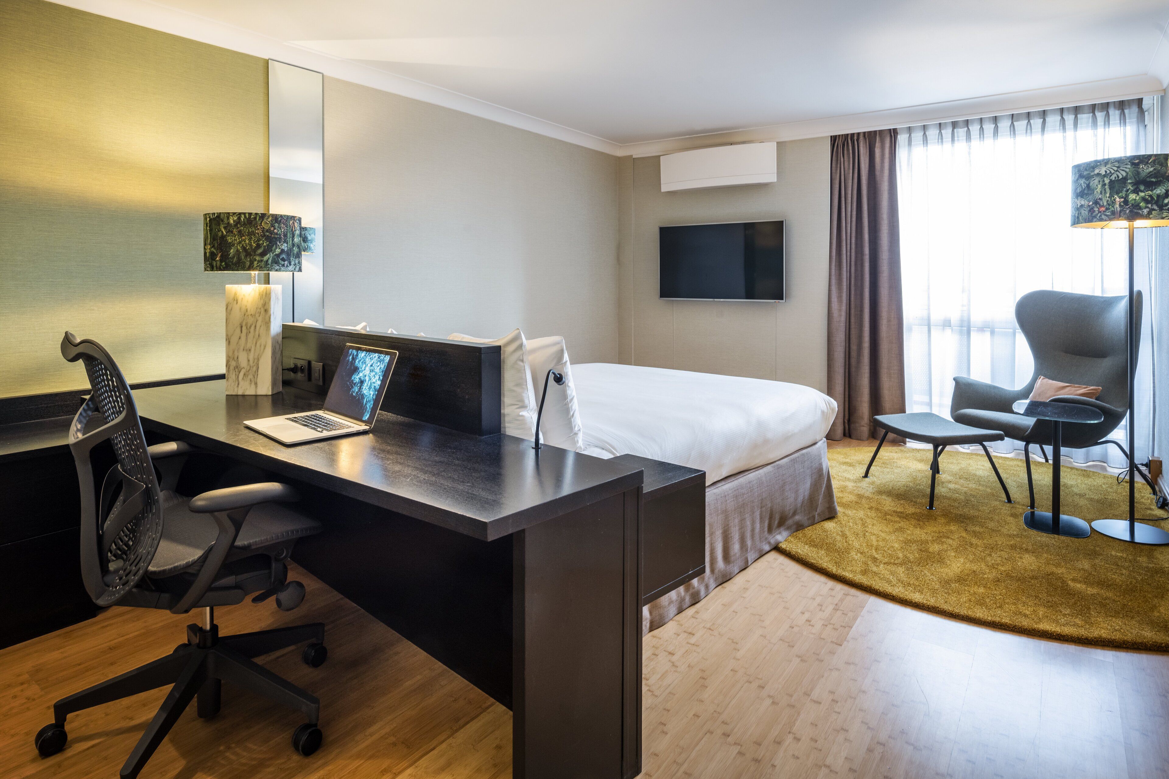 A hotel room with a bed, desk, chair, TV, and a large window with curtains.