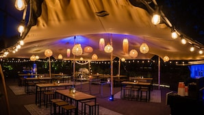 An outdoor dining area with tables and chairs under a large tent, string lights, and hanging lanterns.