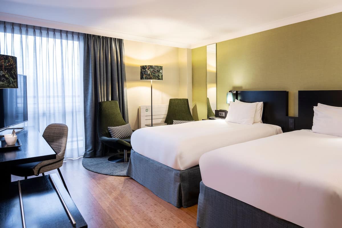 A hotel room with two beds, a desk, a chair, and a TV.