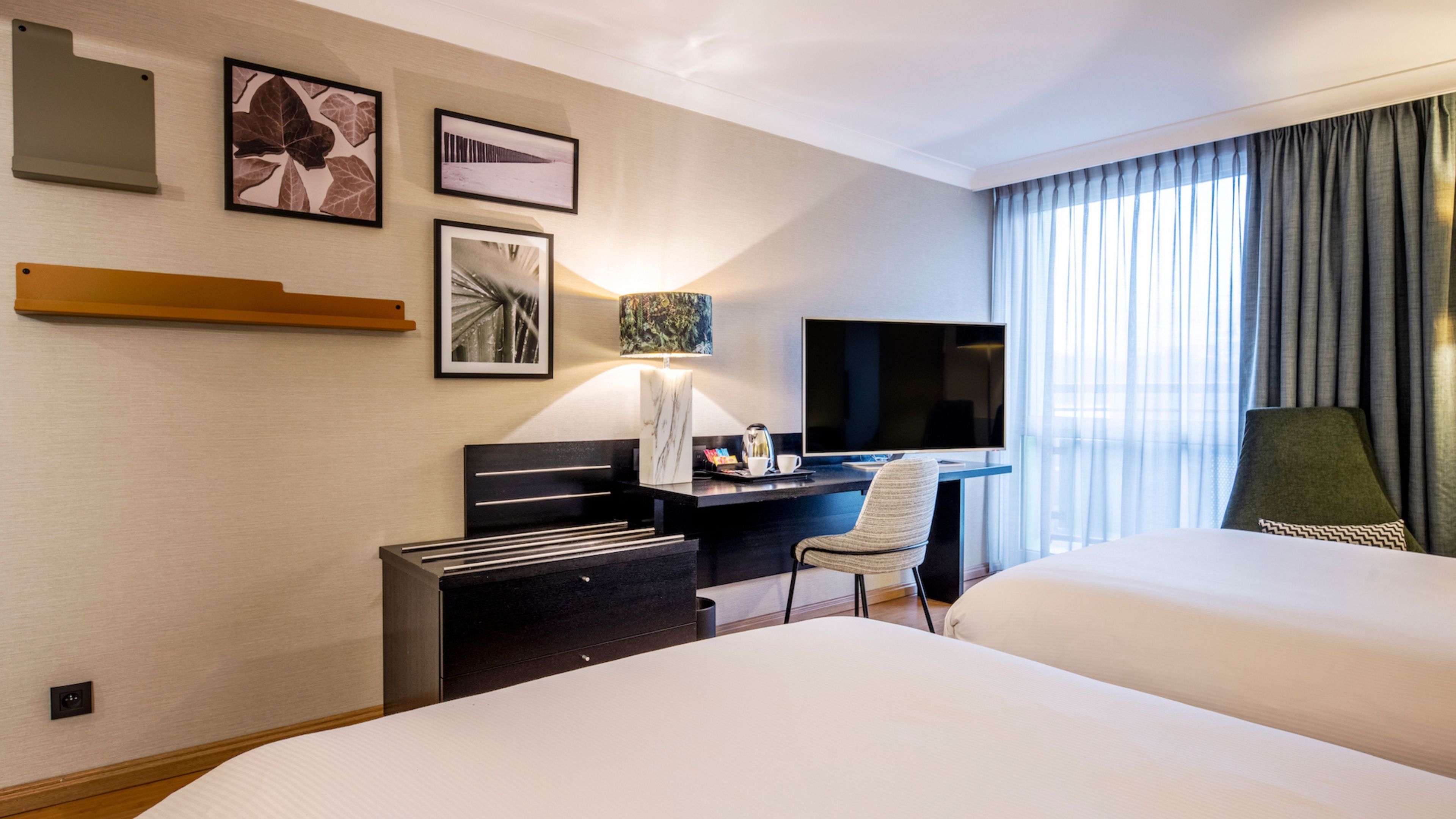 A hotel room with two beds, a desk with a flat-screen TV, a lamp, and framed pictures on the wall.