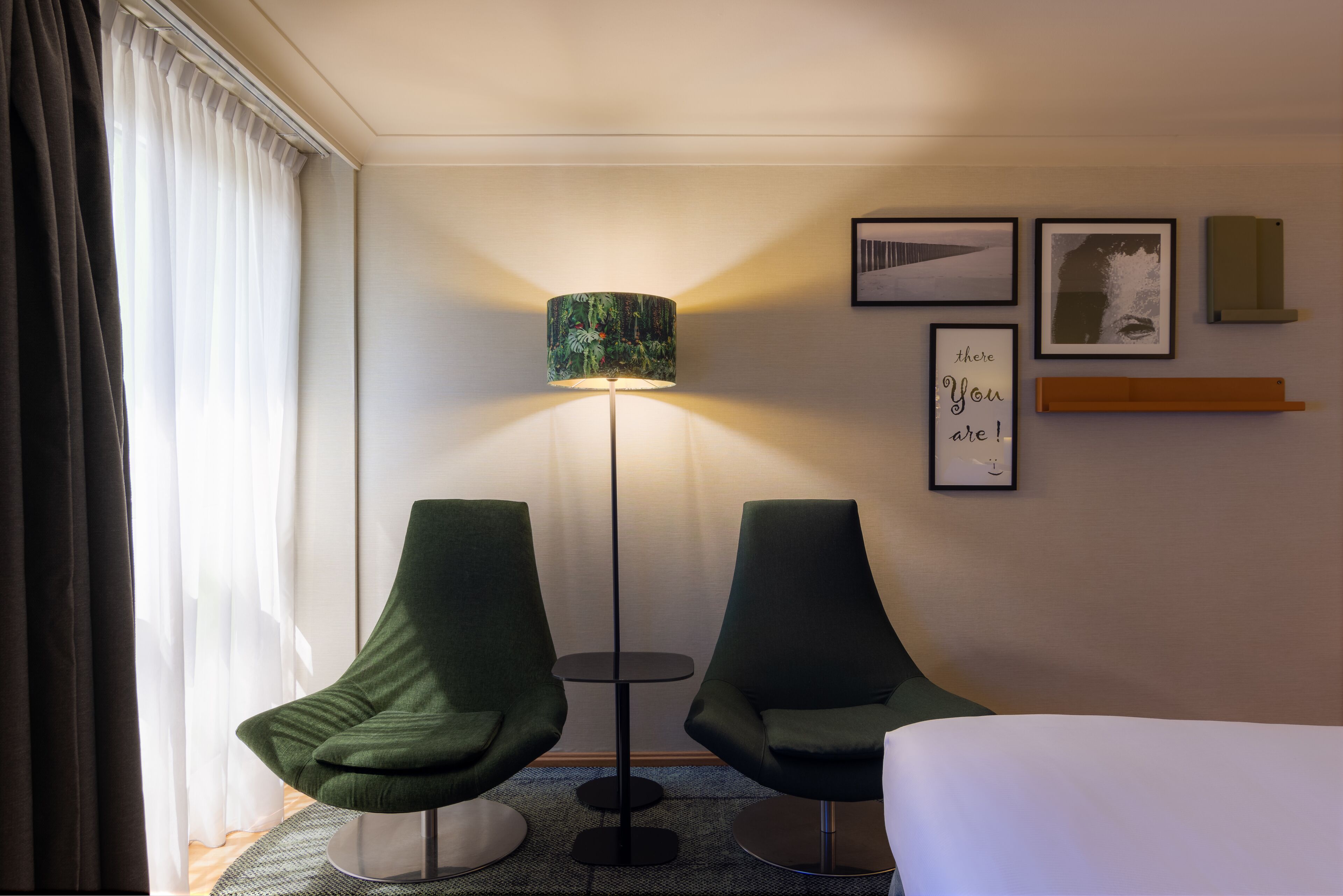 Une chambre d’hôtel avec deux fauteuils verts, une lampe sur pied, un lit et une petite table. Des tableaux encadrés ornent le mur, et une étagère contient un livre.