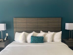 A neatly made bed with white linens and a teal pillow, a wooden headboard, and bedside lamps on nightstands. - Baymont by Wyndham Lumberton NC (Lumberton)