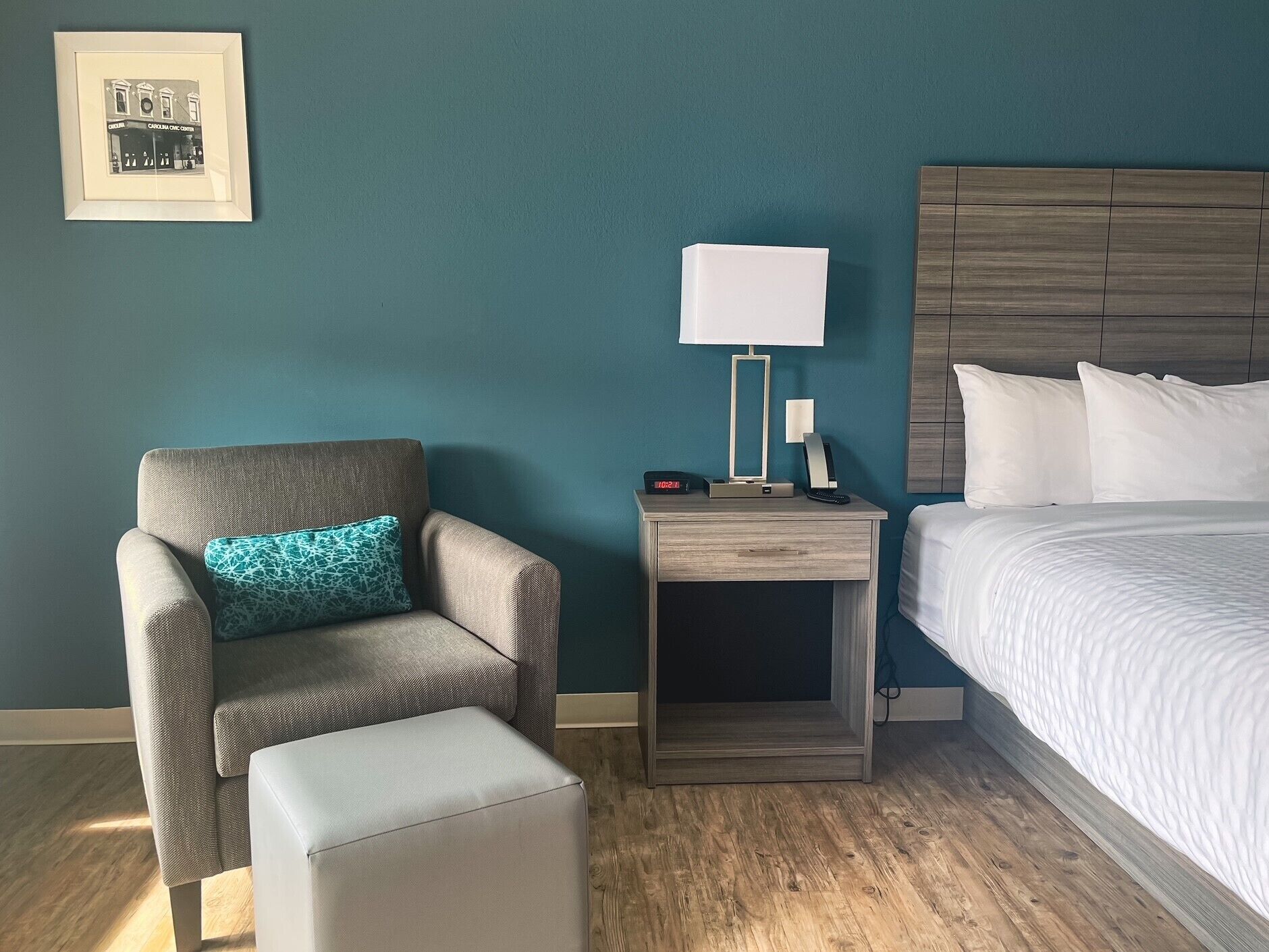 Ein Hotelzimmer mit einem Bett, einem Nachttisch mit Lampe, einem Sessel mit Kissen und einem gerahmten Bild an der Wand.