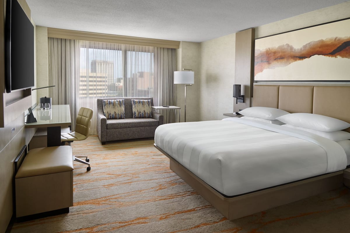 A hotel room with a large bed, a desk with a chair, a sofa, a TV, and a painting on the wall.