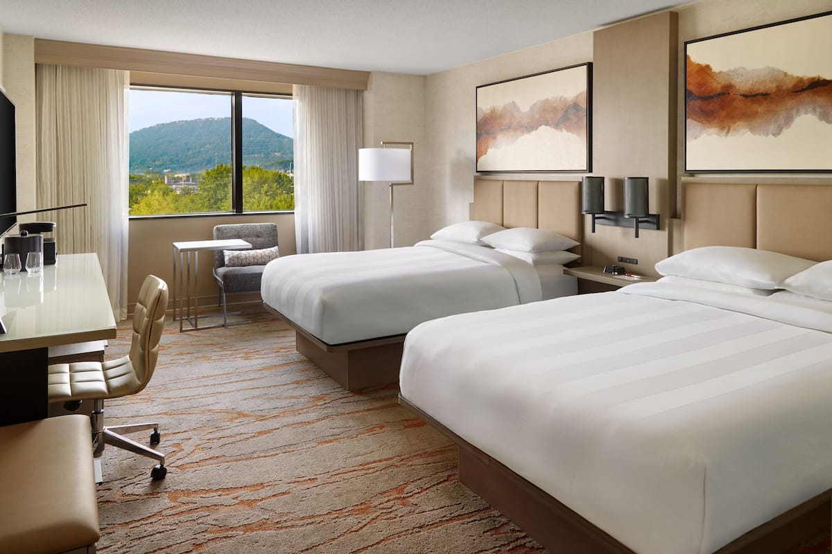 A hotel room with two beds, a desk, a chair, a lamp, and a view of the mountains.