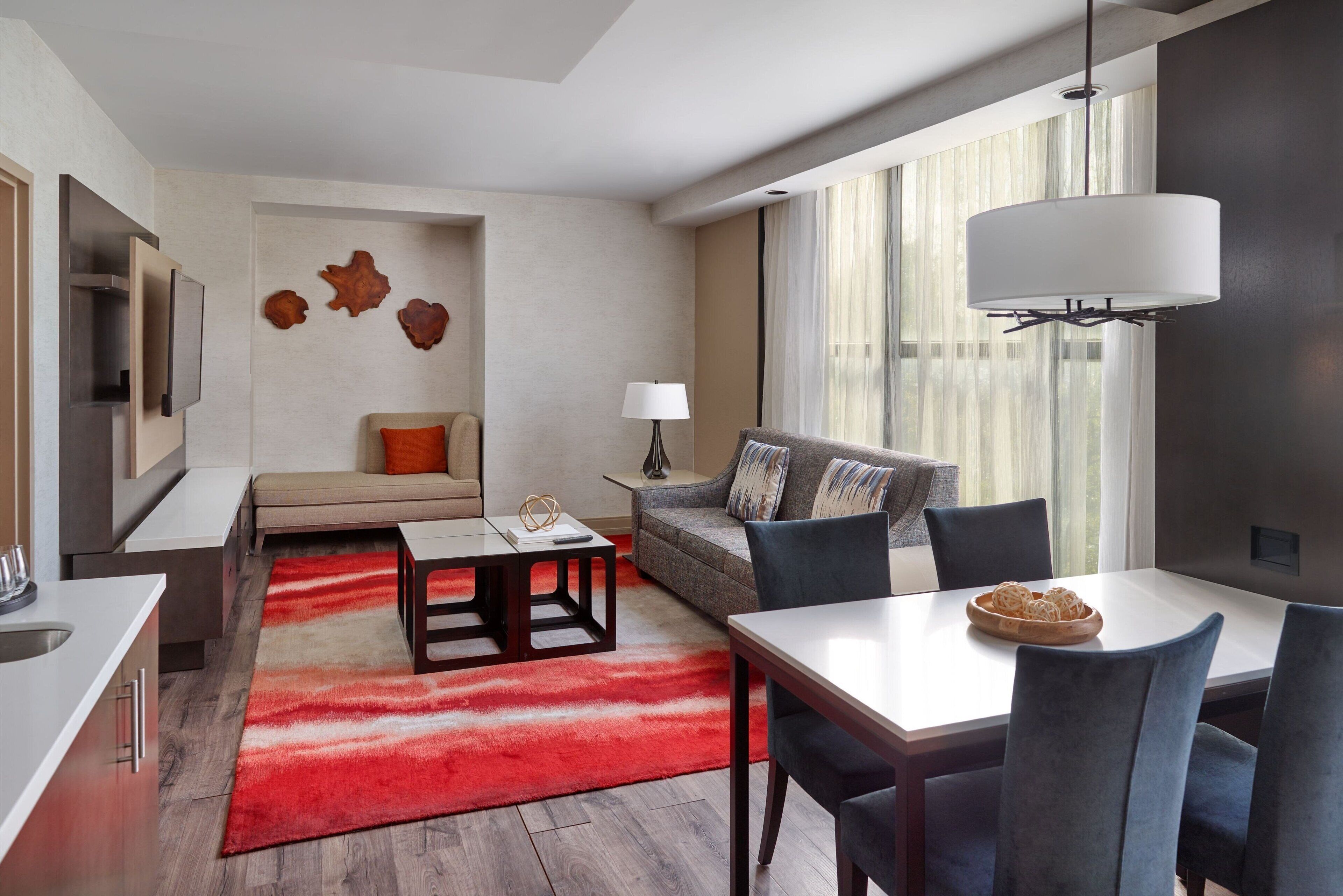 Une chambre d’hôtel moderne, dotée d’un canapé, d’une table à manger et de chaises, d’un tapis rouge et d’une grande fenêtre avec des rideaux.