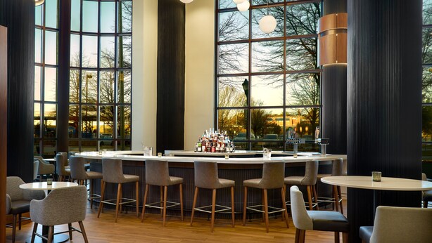 Un área de bar moderna con barra central, diversas opciones de asientos y grandes ventanales que ofrecen vistas al exterior.
