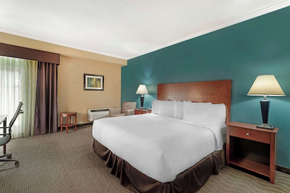 A hotel room with a large bed, two bedside tables with lamps, a chair, a small table, and a window with curtains.