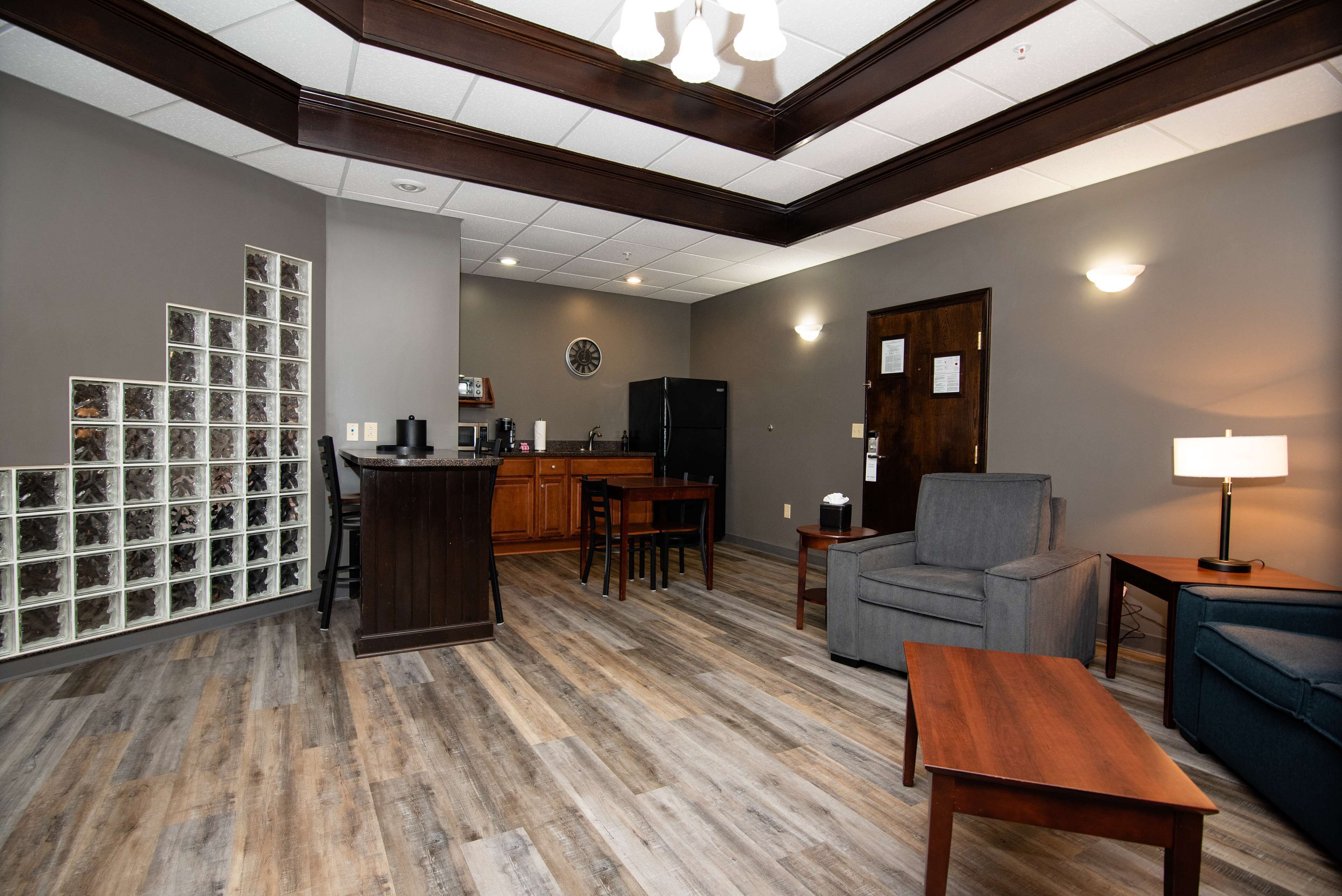 A modern living room with a wooden floor, a grey sofa, a wooden coffee table, a kitchen area with a microwave, and a refrigerator.