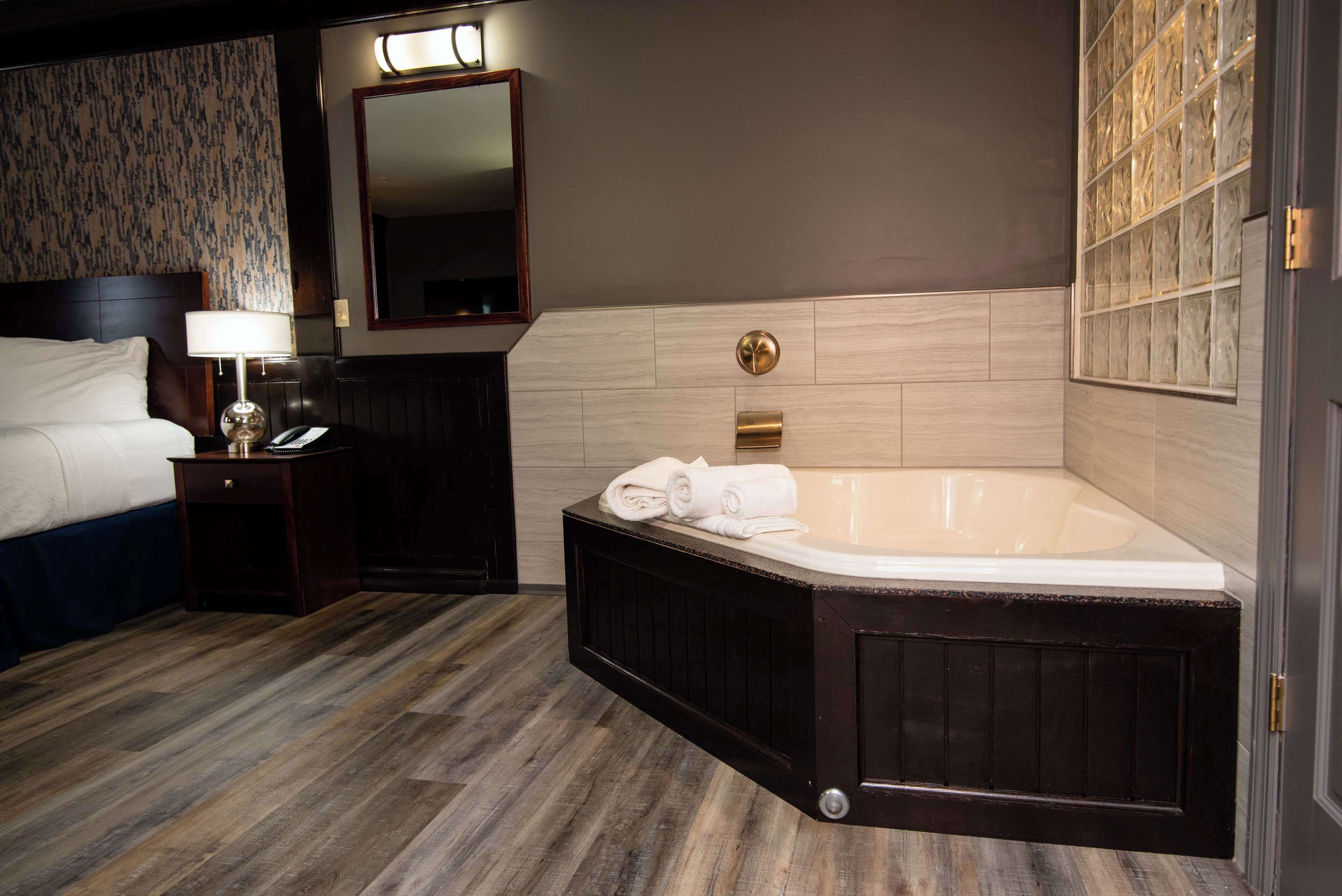 A hotel room with a large bathtub, a bedside table with a lamp, a mirror, and a patterned wall.