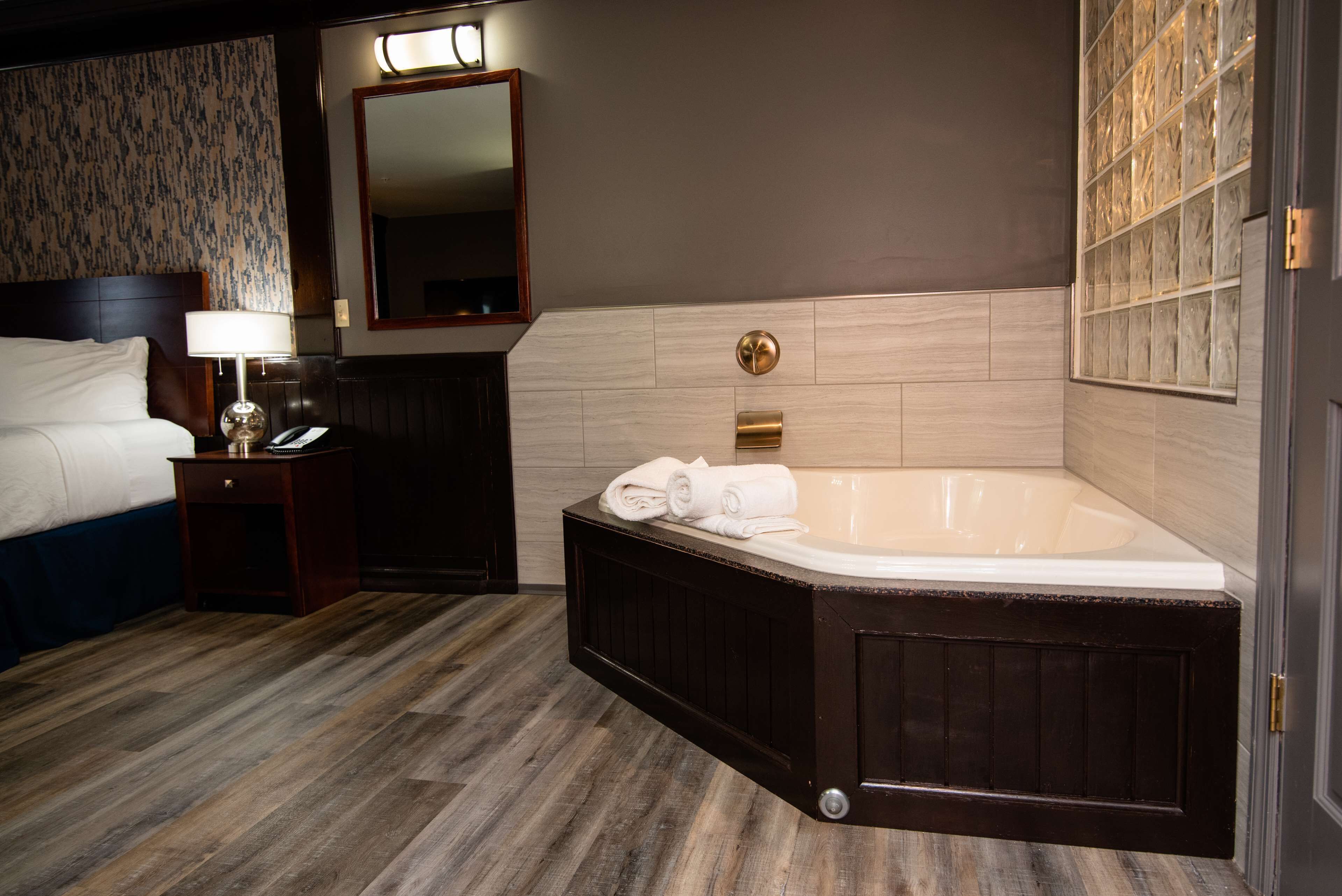 Habitación de hotel con una cama grande, baño separado con bañera, piso de madera y un espejo sobre la cama.