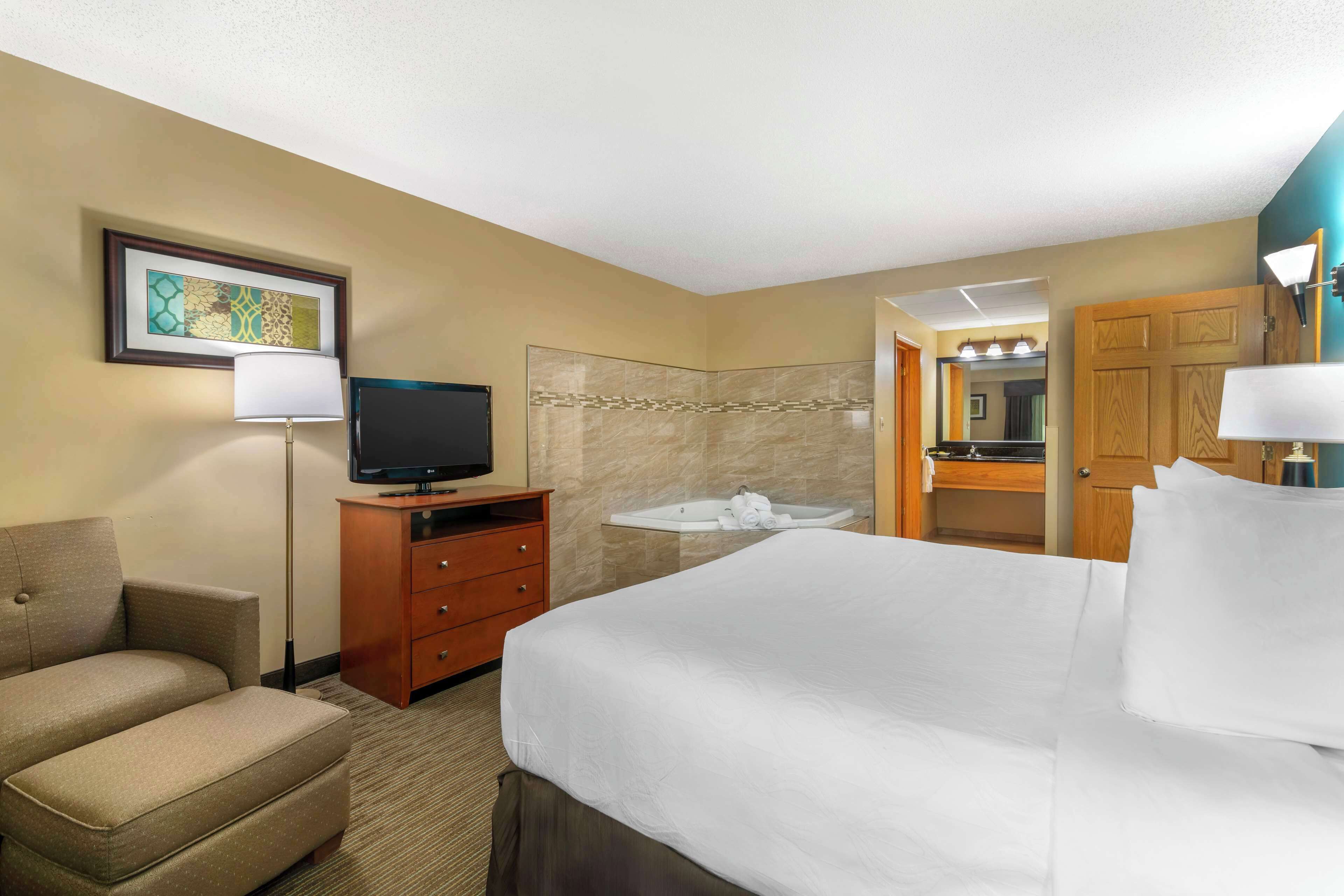 Habitación de hotel con cama, televisor, sofá, escritorio con lámpara, baño con lavabo y espejo, y un cuadro en la pared.