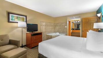 A hotel room with a bed, a TV, a sofa, a desk with a lamp, a bathroom with a sink and mirror, and a framed picture on the wall.
