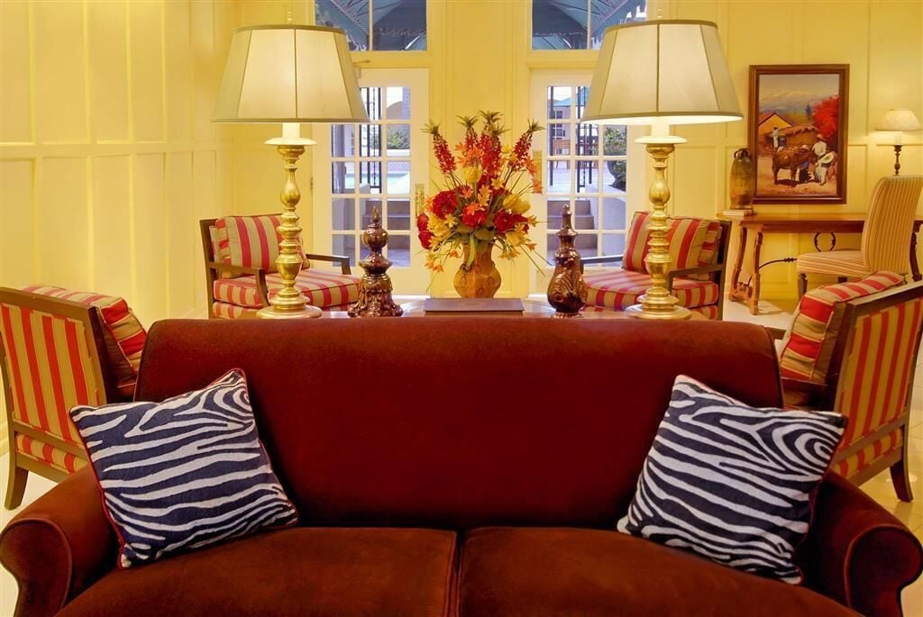 A red sofa with zebra-patterned pillows, two lamps, a vase with flowers, and striped chairs in a warmly lit room with large windows.