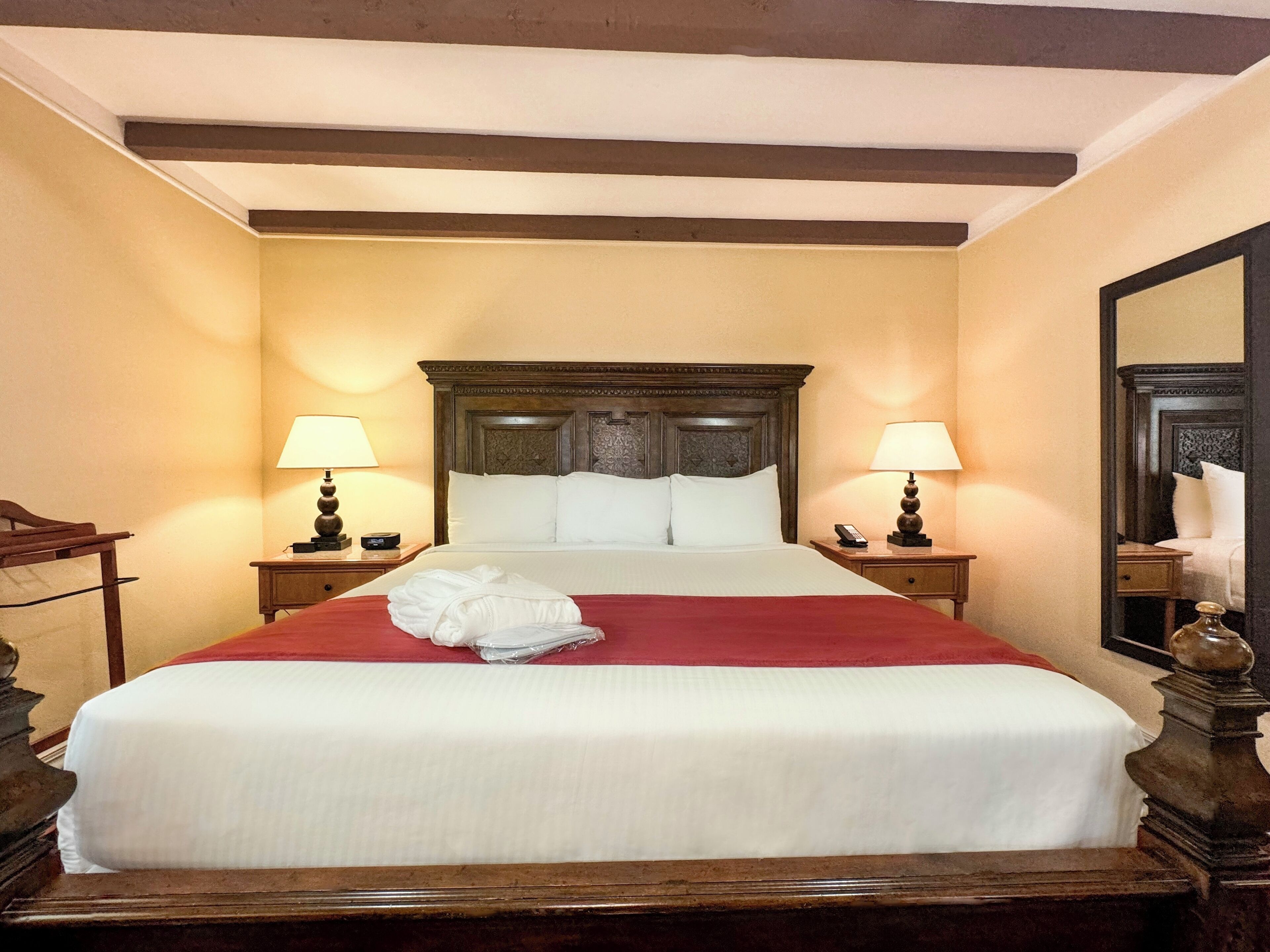A hotel room with a large bed, two bedside tables with lamps, a wooden headboard, and a mirror on the wall.
