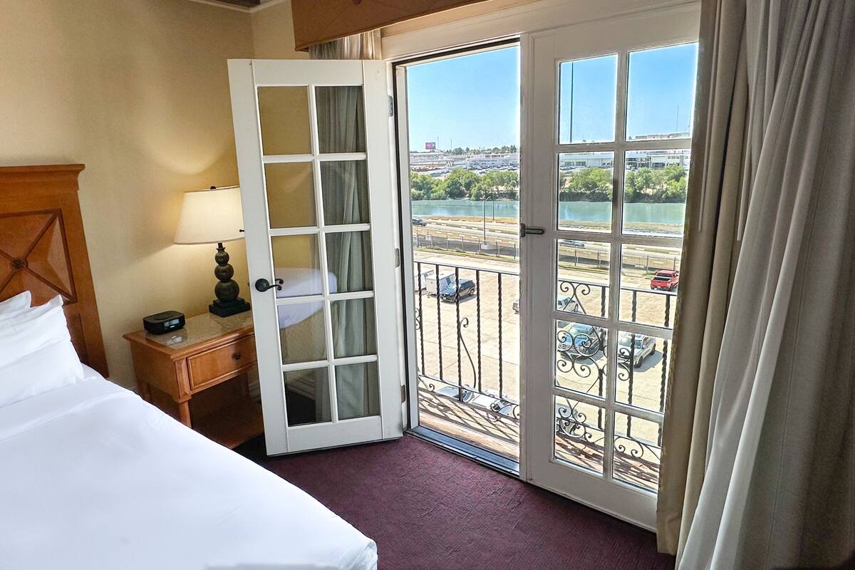 A hotel room with a bed, a nightstand, a lamp, and a view of a balcony with a railing and a parking lot.