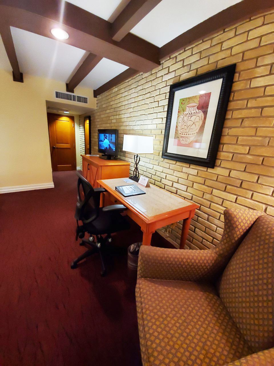 A hotel room with a brick wall, a television, a desk with a chair, and a framed picture.