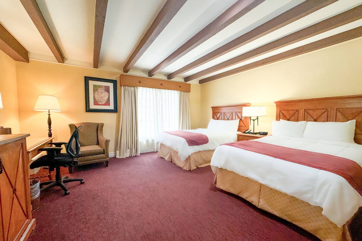 A hotel room with two beds, a desk, a chair, and a lamp.