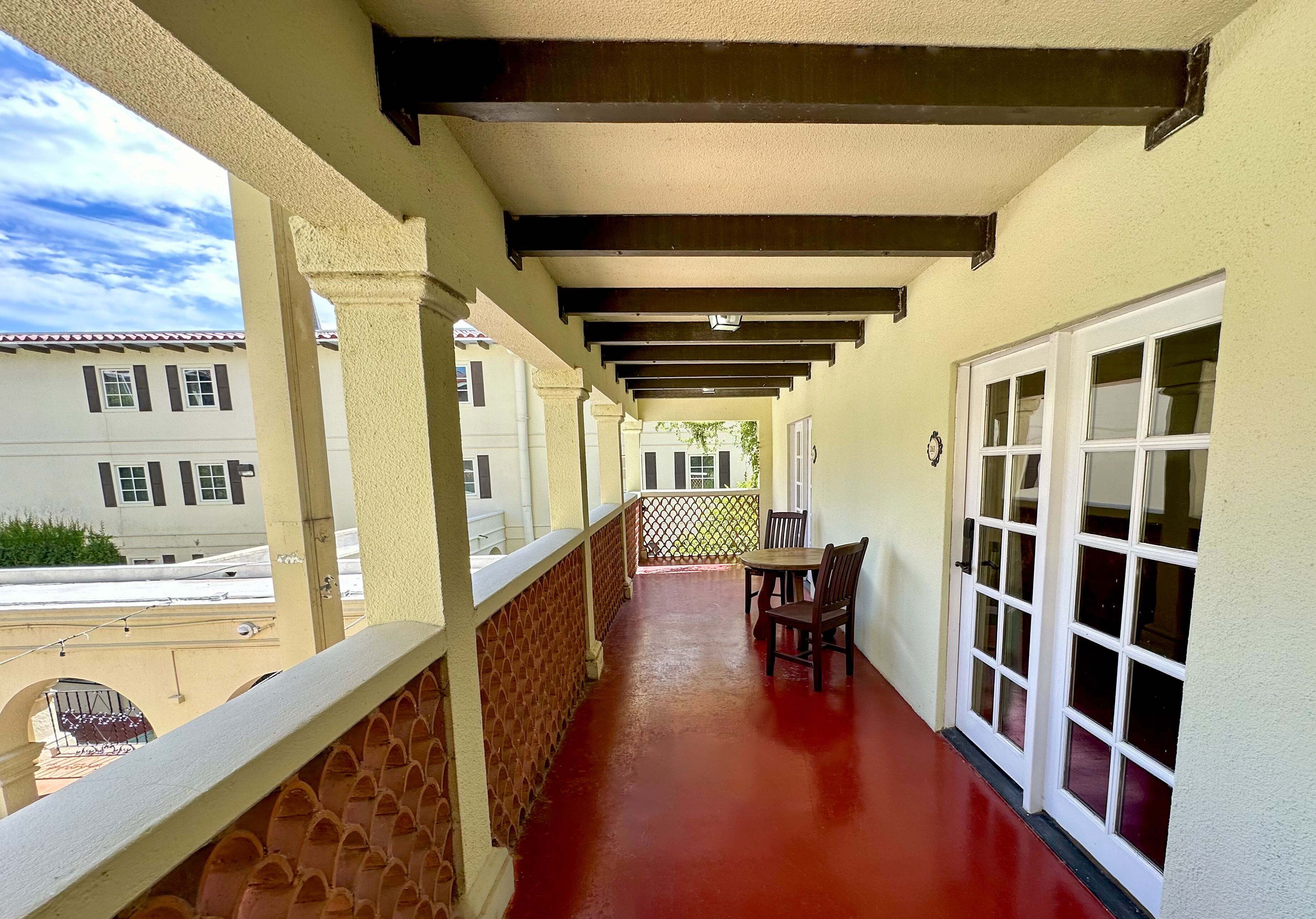 Un balcón techado con piso rojo, barandas blancas y una mesa con sillas.