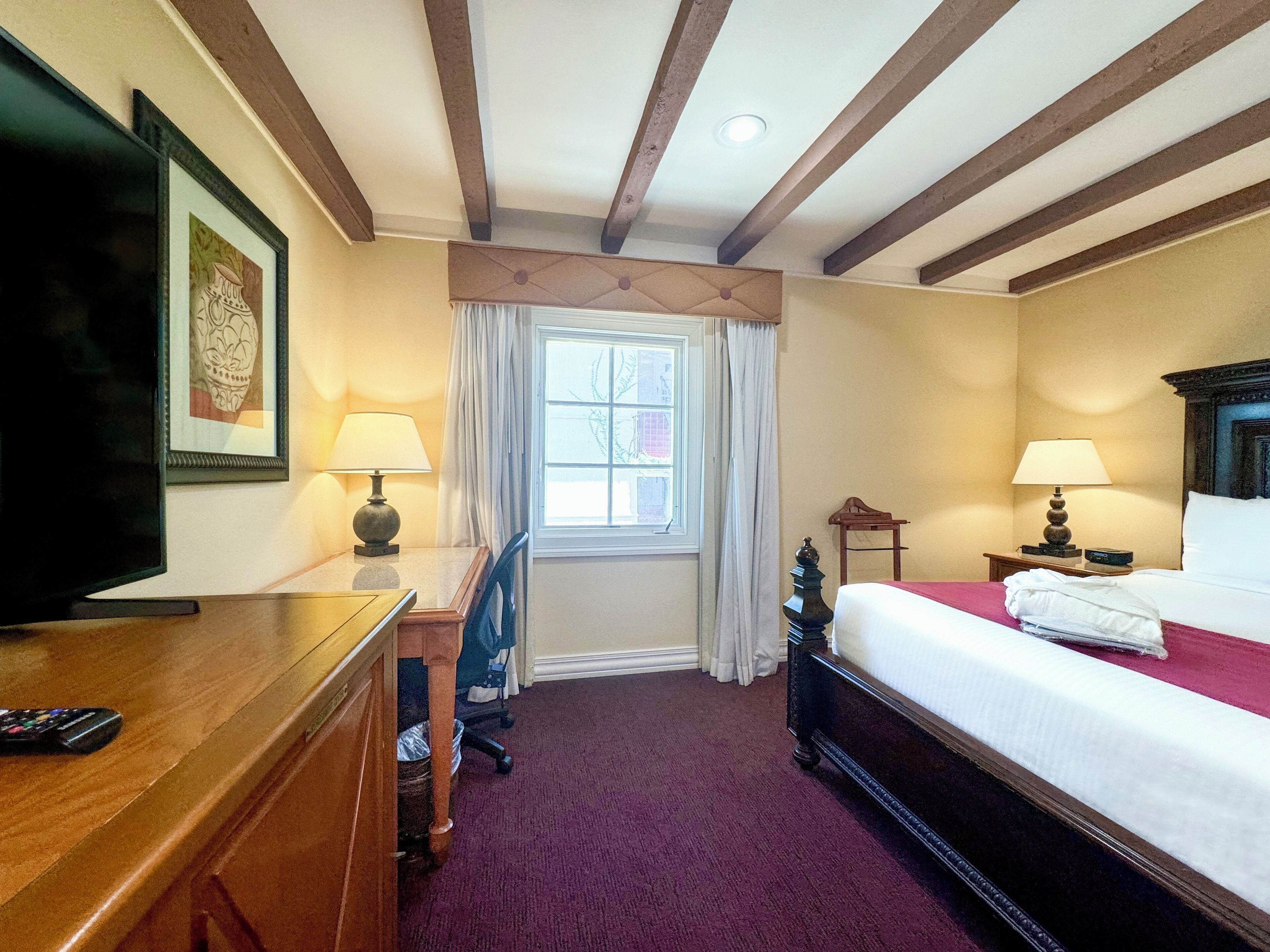 A hotel room with a bed, a desk with a lamp, a television, and a window with curtains.
