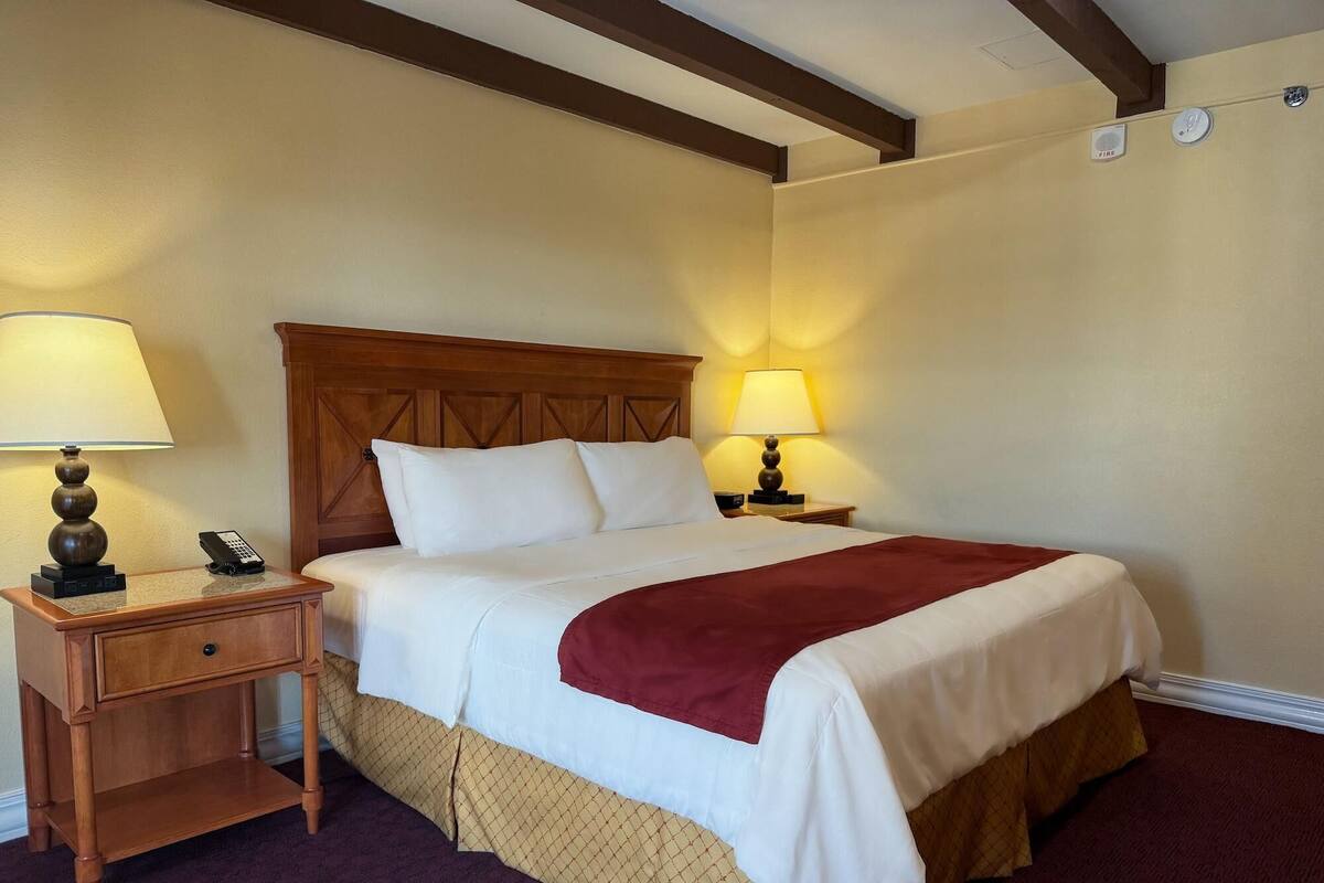 A hotel room with a wooden headboard, two bedside lamps, a telephone, a bed with white bedding and a red throw, and a carpeted floor.