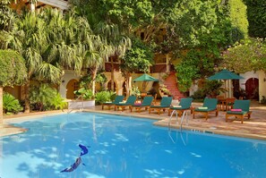 A pool area with lounge chairs and umbrellas, surrounded by lush greenery and a building with arches.