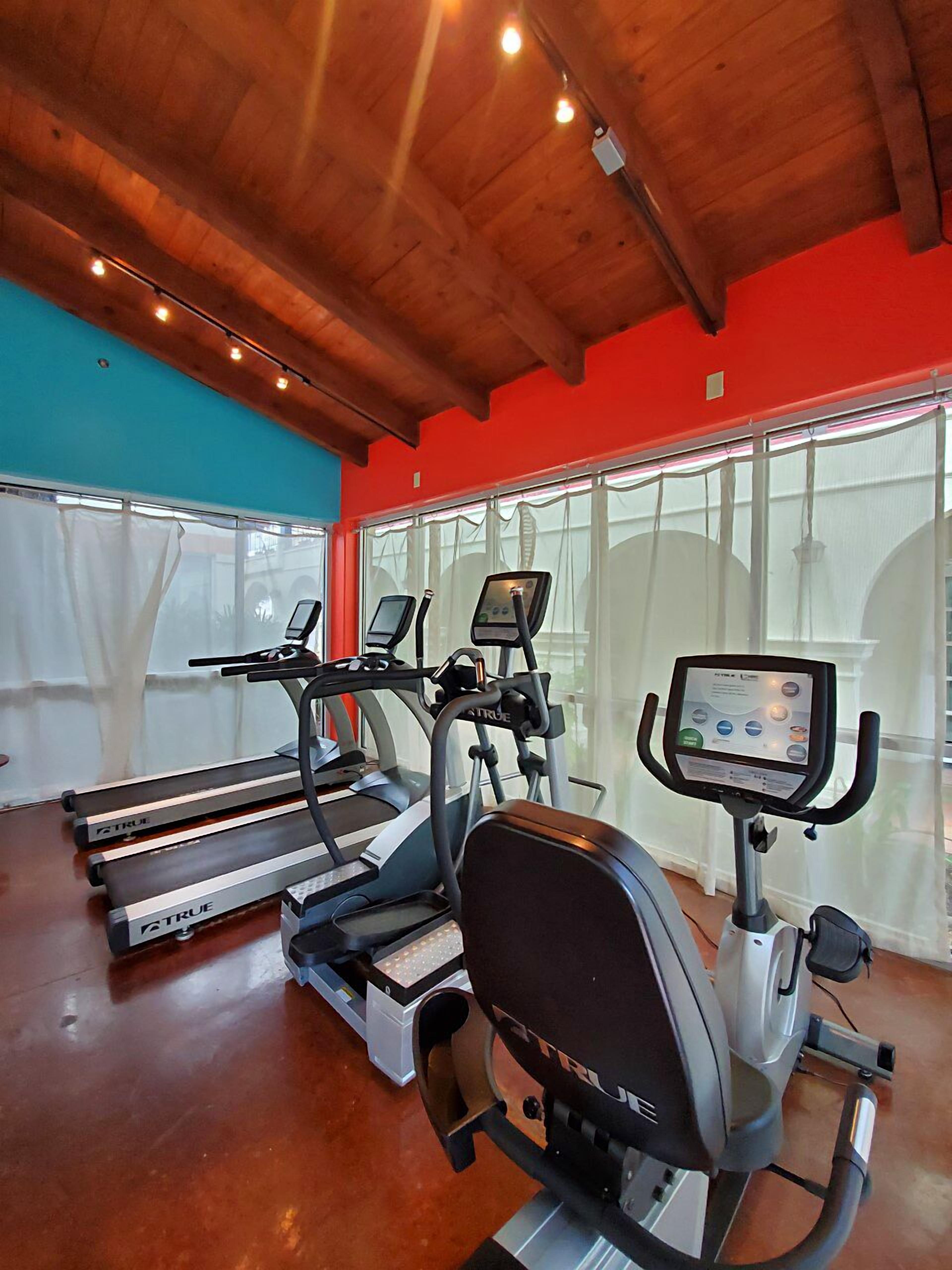 A gym with treadmills, an elliptical, and a stationary bike, featuring a wooden ceiling and large windows.