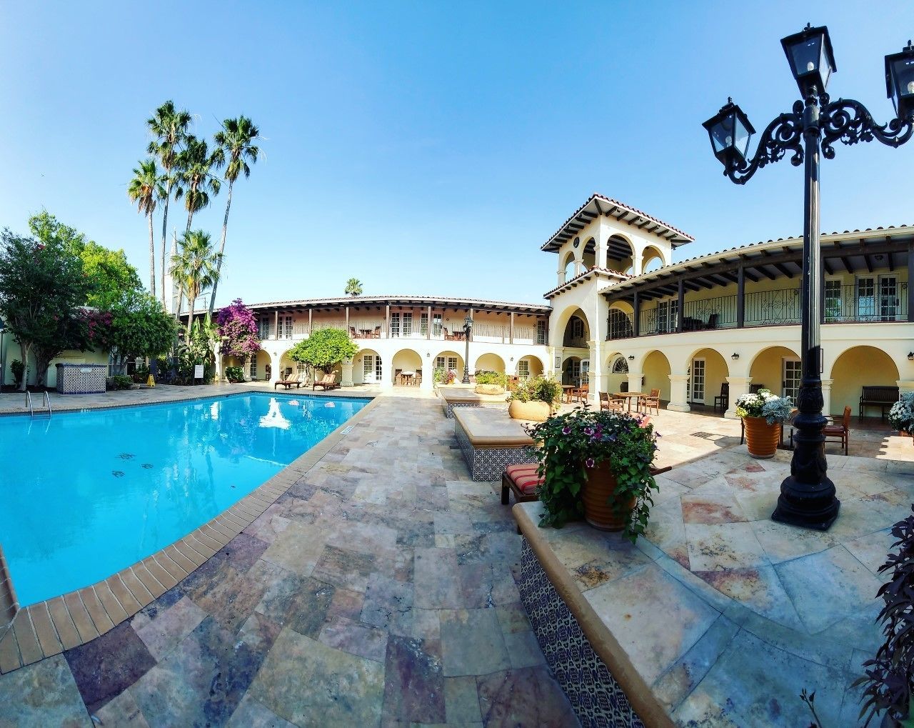 A pool area with a clear blue pool, surrounded by a tiled patio, outdoor seating, and a building with arches and a bell tower.