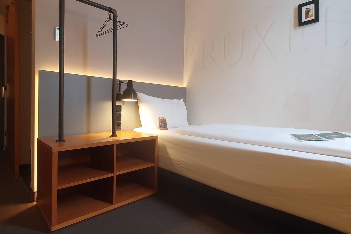 Une chambre d’hôtel avec un lit simple, une table de chevet avec une lampe, une étagère et un porte-serviettes fixé au mur.