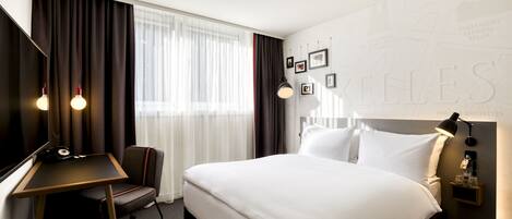 A hotel room with a large bed, a desk, a chair, a lamp, and a wall with framed pictures.