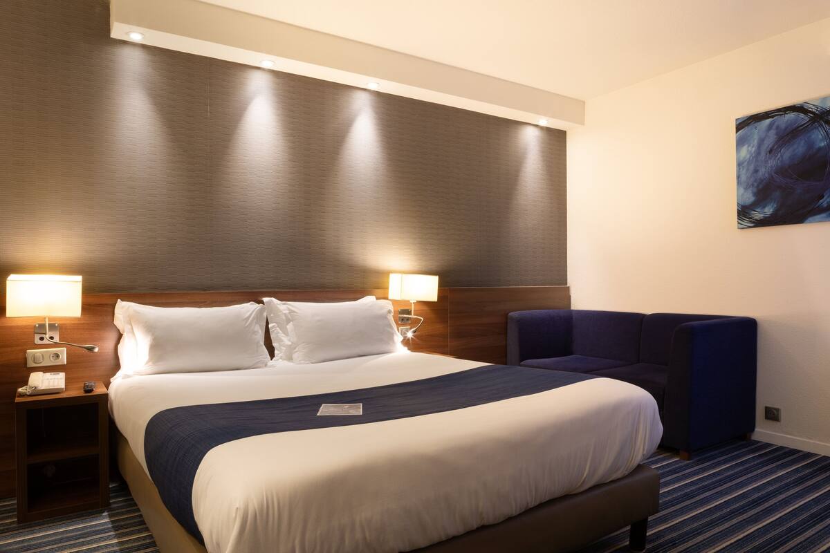 A hotel room with a large bed, two bedside lamps, a nightstand, a blue armchair, and a painting on the wall.