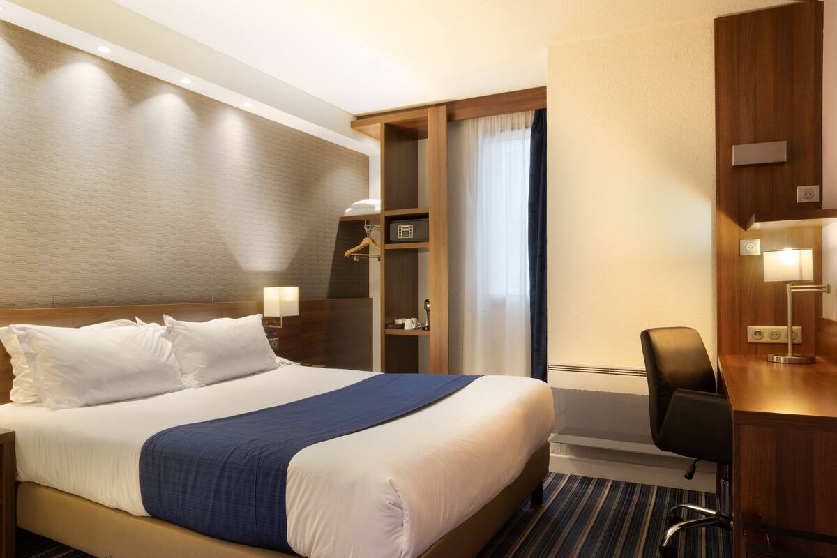 A hotel room with a large bed, bedside tables, a desk with a chair, and a wardrobe.