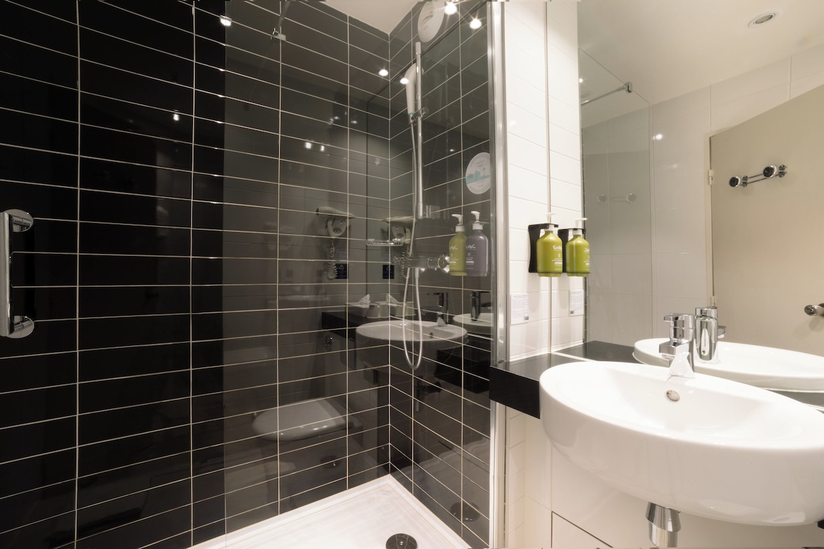 Une salle de bain moderne avec des murs recouverts de carreaux noirs, une douche en verre et deux vasques blanches dotées de robinetterie chromée.