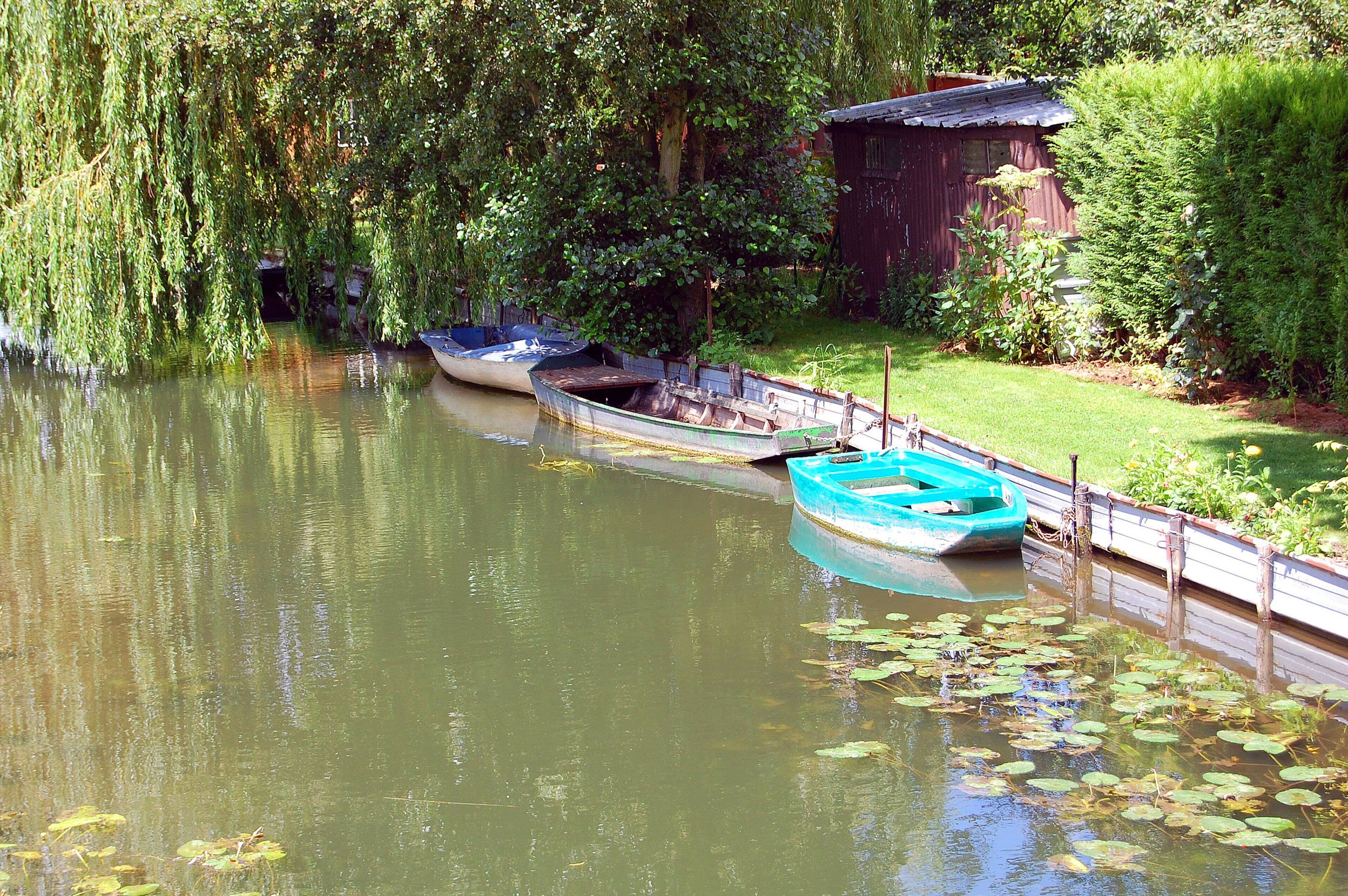 A calm river with boats docked along the bank, surrounded by lush greenery and a small wooden structure.