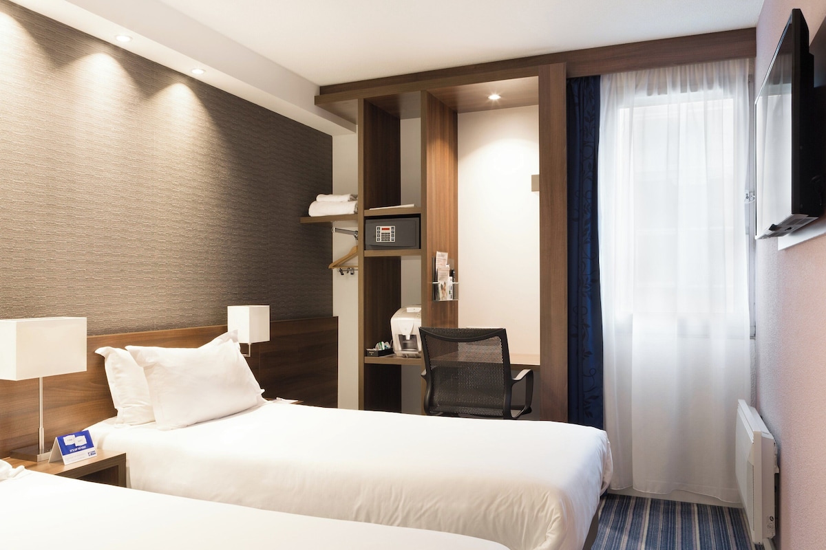 Une chambre d’hôtel avec deux lits, un bureau, une chaise, une télévision et une fenêtre avec des rideaux.