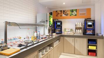 A self-service breakfast area with a coffee machine, milk dispensers, and a variety of food items.