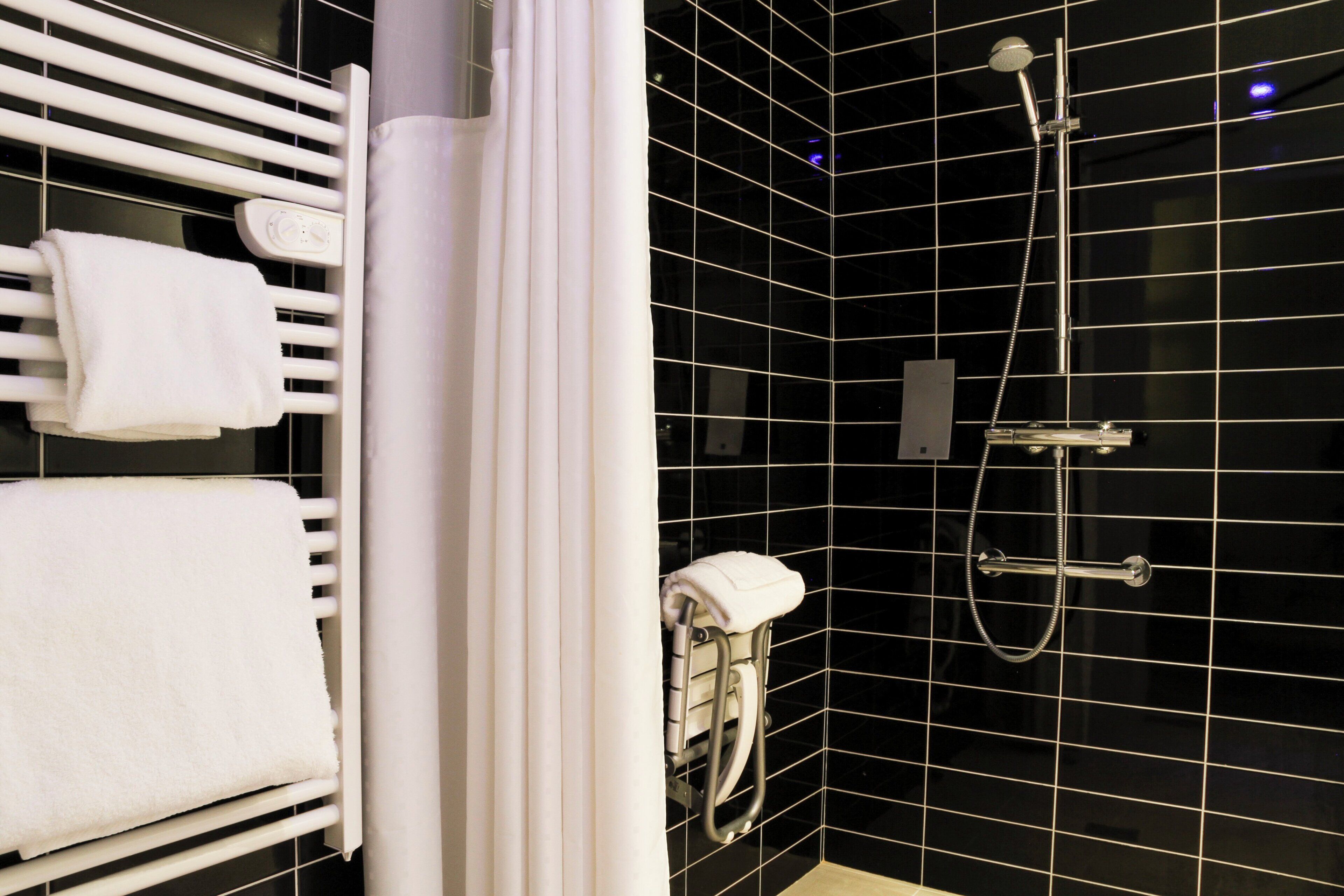 Une salle de bain moderne avec un mur recouvert de carreaux noirs, un porte-serviettes blanc, une douche équipée d’un pommeau de douche à main et des toilettes dotées d’un support pour le papier essuie-tout.
