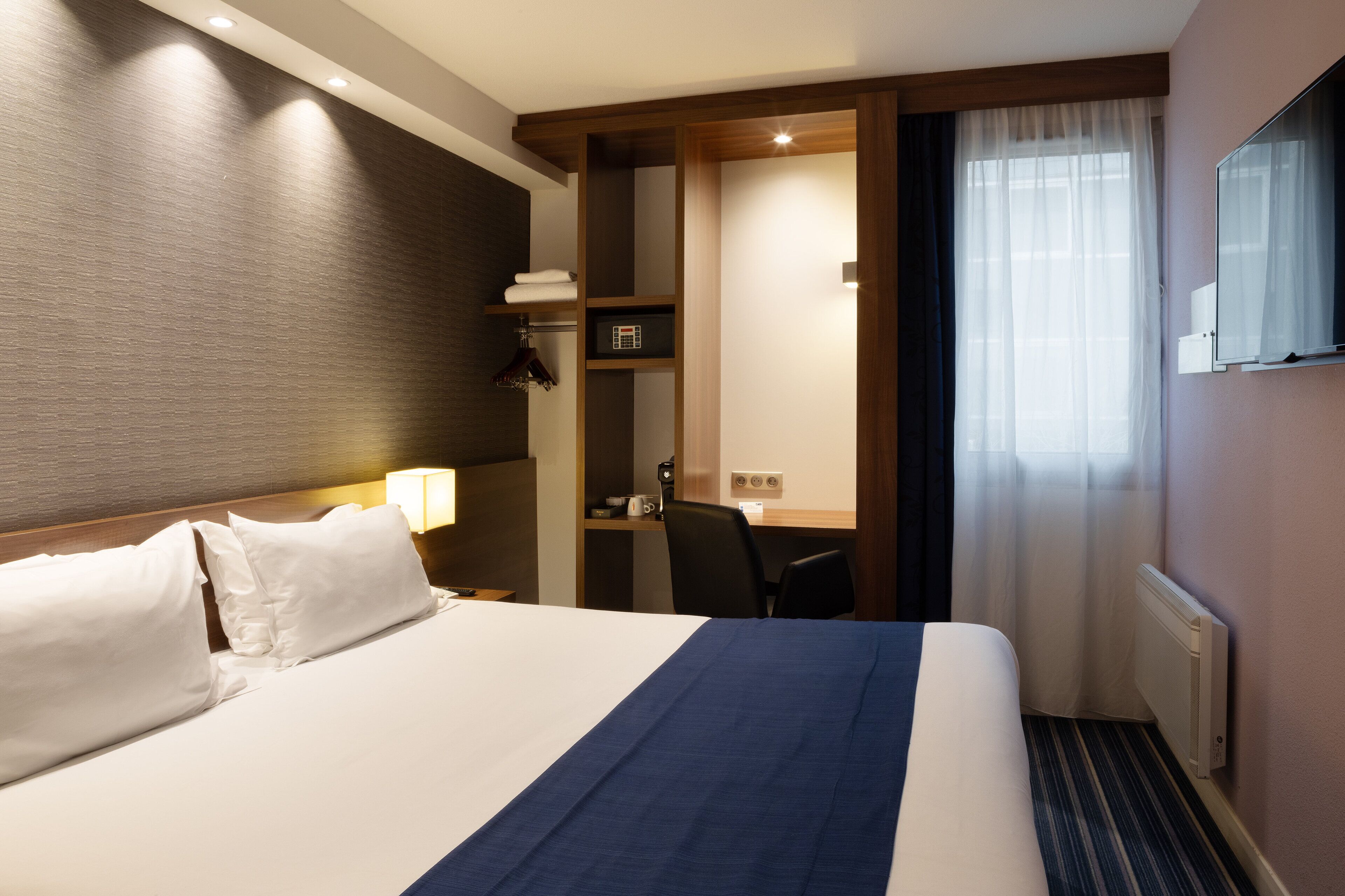A hotel room with a large bed, a desk, a chair, a wardrobe, and a window with curtains.