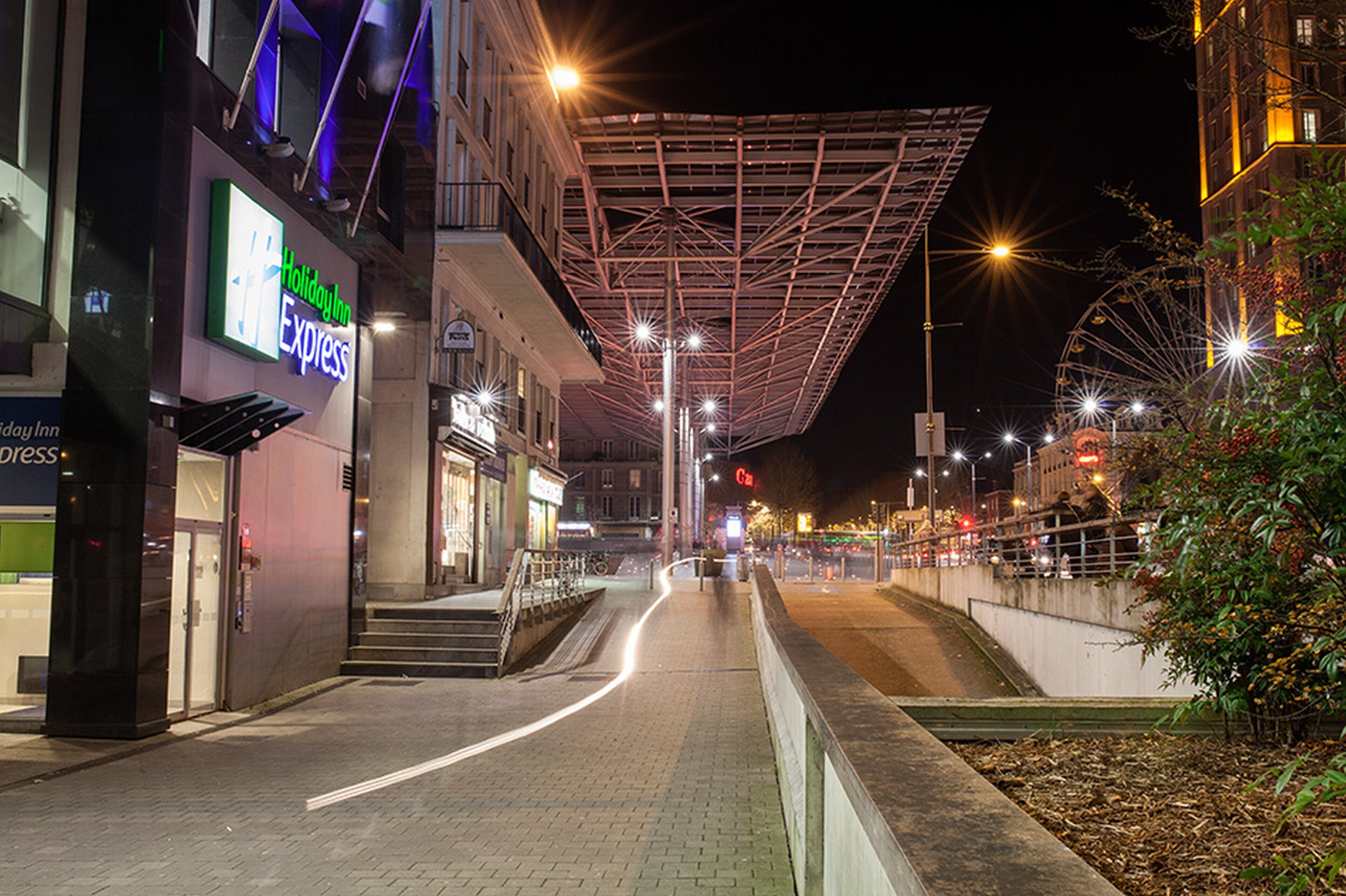 Eine beleuchtete Stadtstraße bei Nacht mit einem modernen Gebäude, das eine markante Dachkonstruktion und eine leuchtende Beschilderung für ein Holiday Inn Express aufweist.
