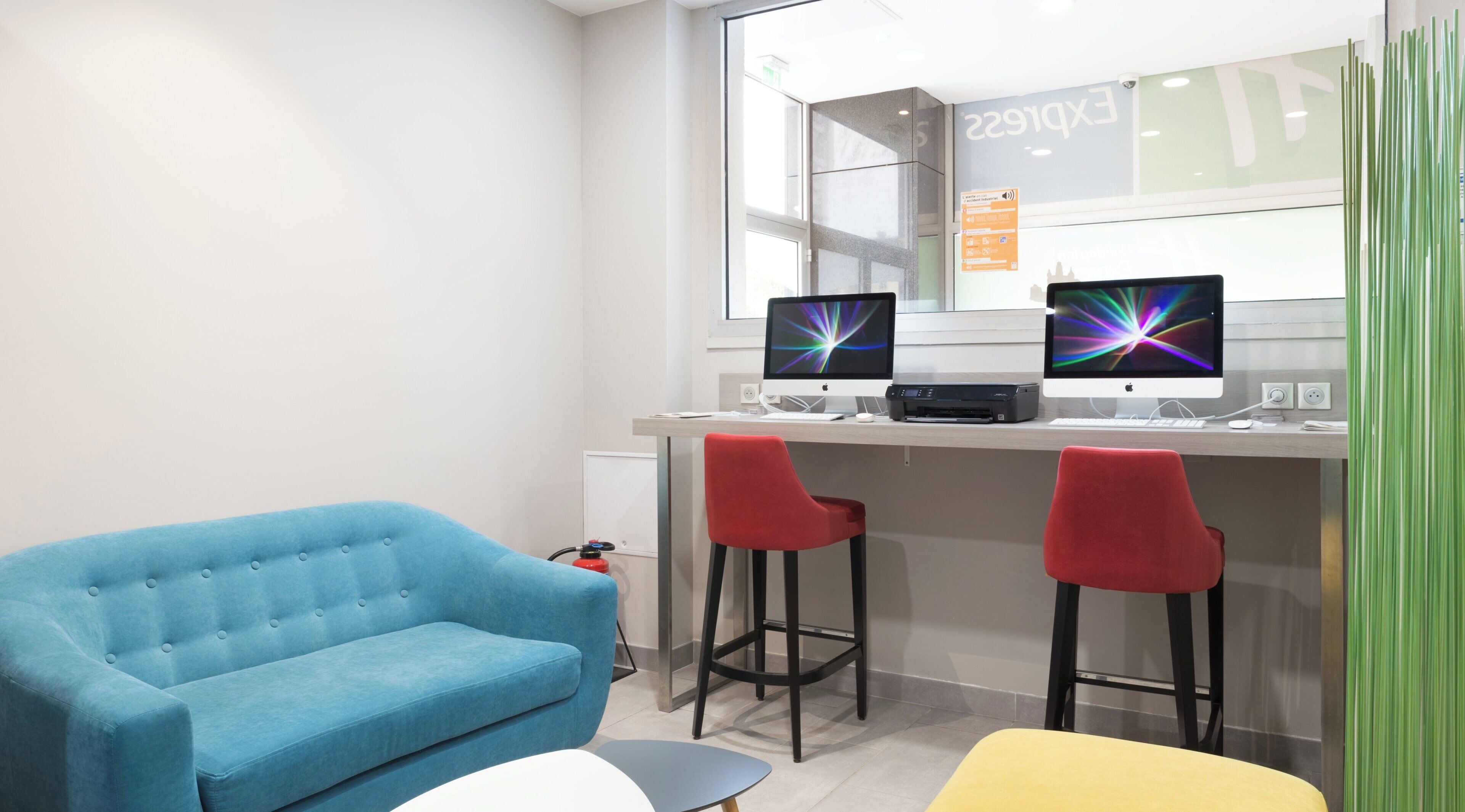 A modern office space with two Apple iMac computers, a blue sofa, two red bar stools, and a yellow chair.