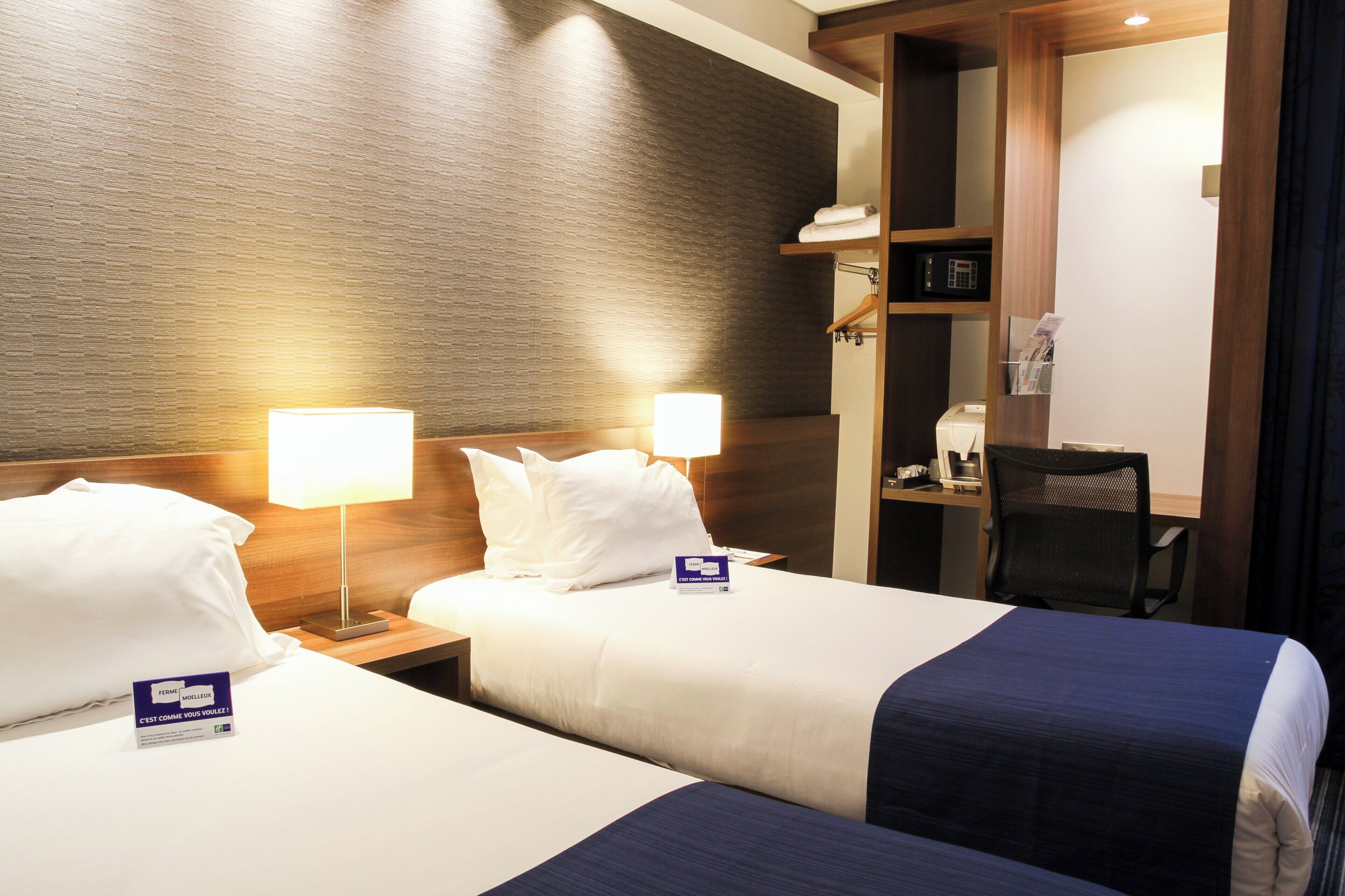 A hotel room with two beds, each with a pillow and a nightstand with a lamp. There is a desk with a chair and a microwave.
